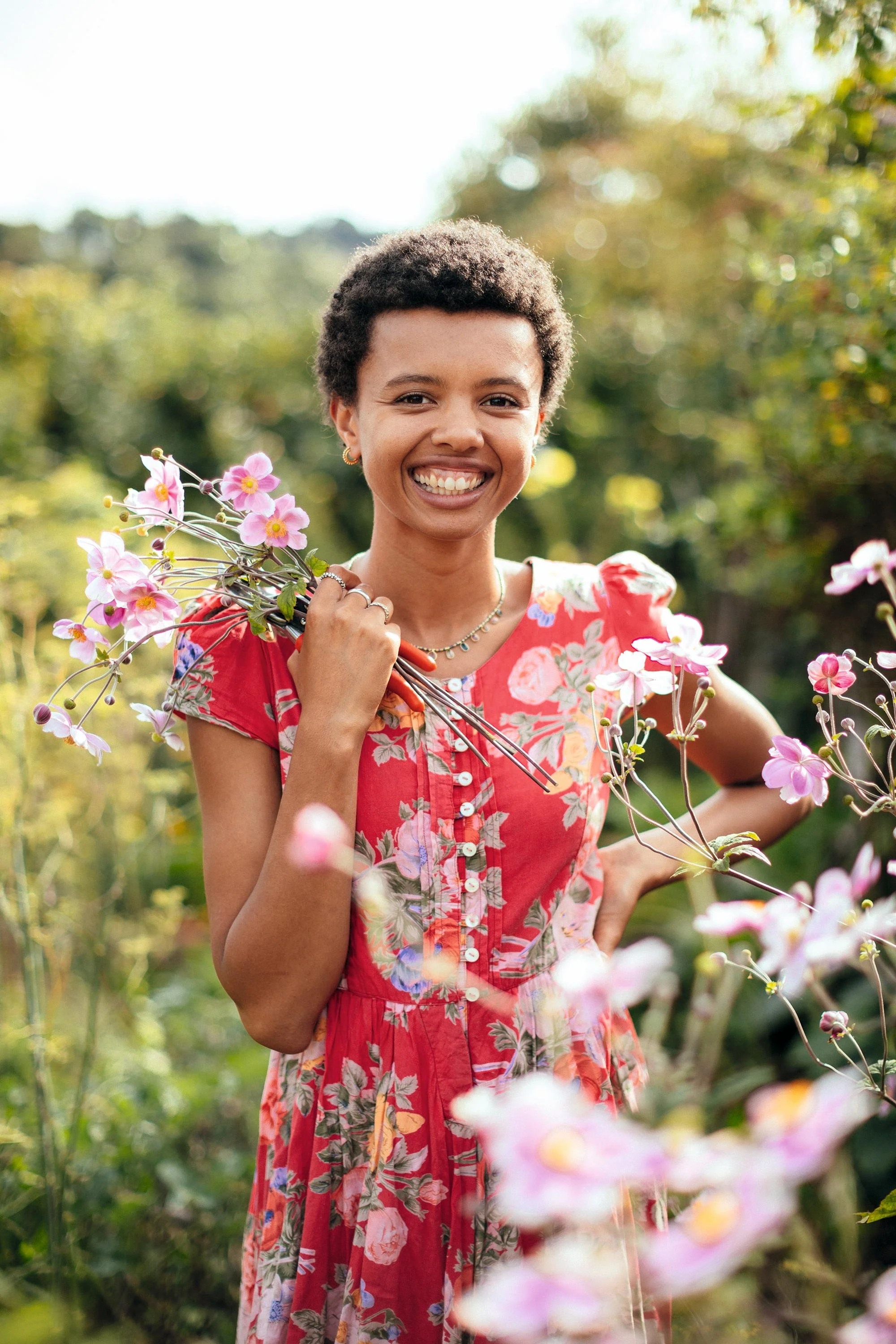 A young woman with short curly hair smiling and holding pink flowers in a garden during daytime.
