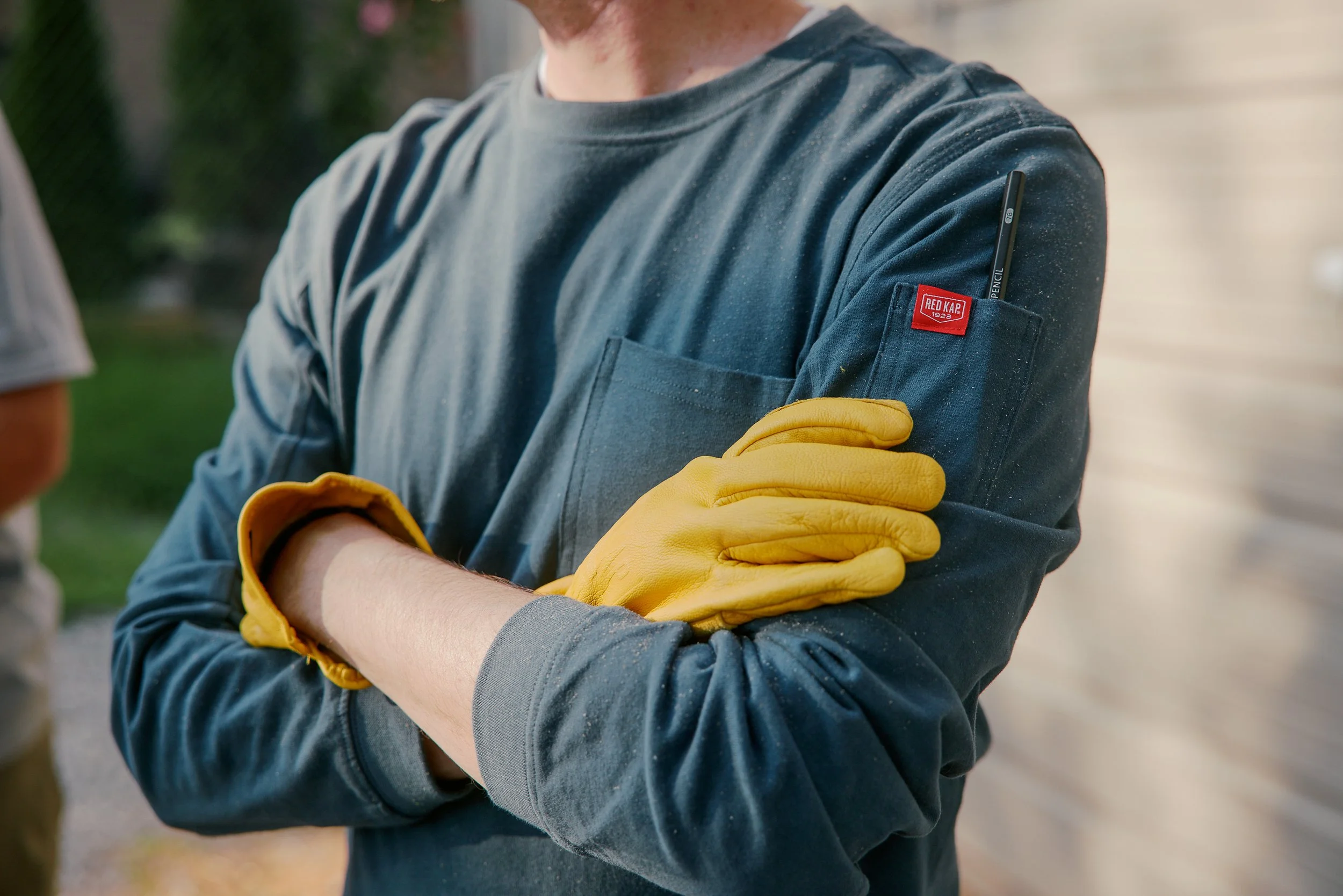 Person wearing a grey long-sleeve shirt with a red logo patch on the sleeve, yellow gloves, crossing their arms, outdoors.