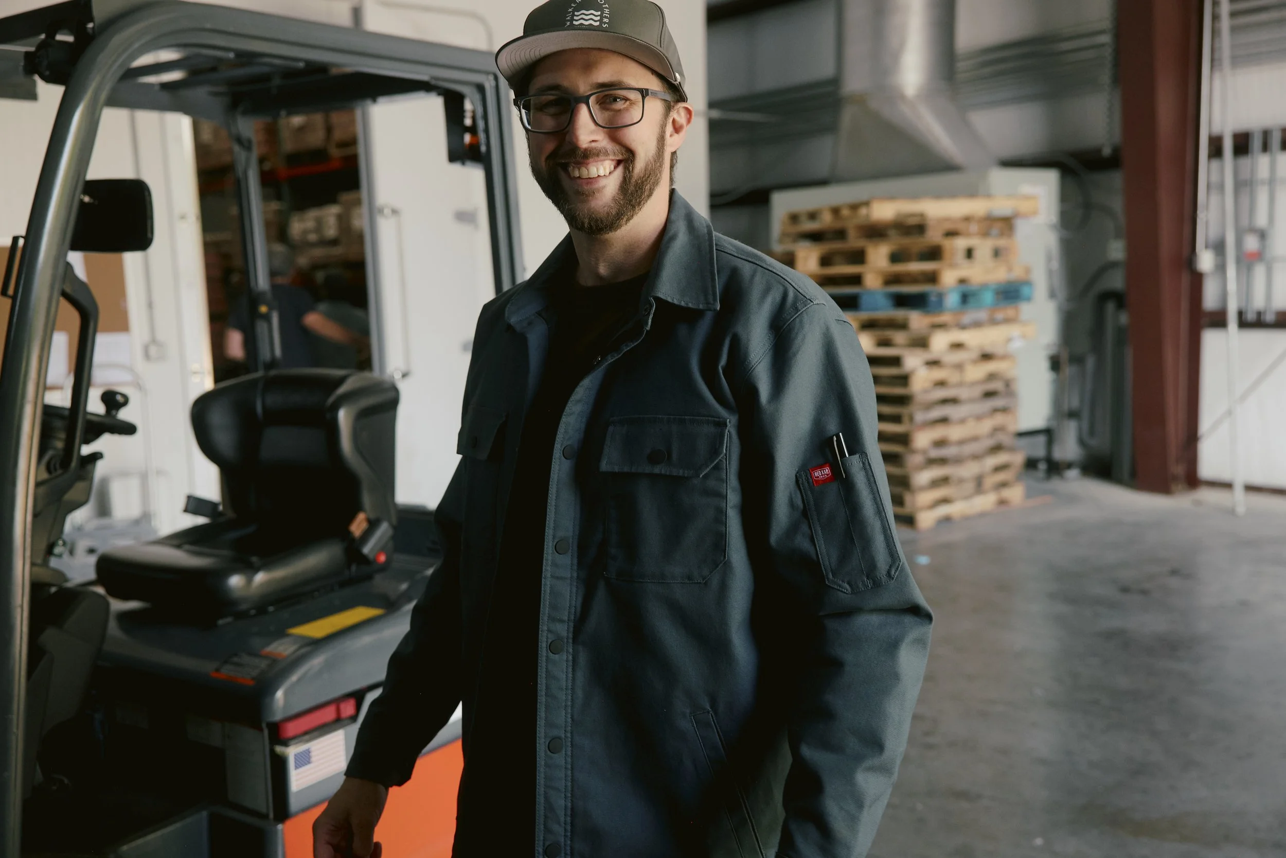 A man wearing glasses, a gray cap, and a dark jacket stands next to a forklift in a warehouse with stacked pallets in the background.