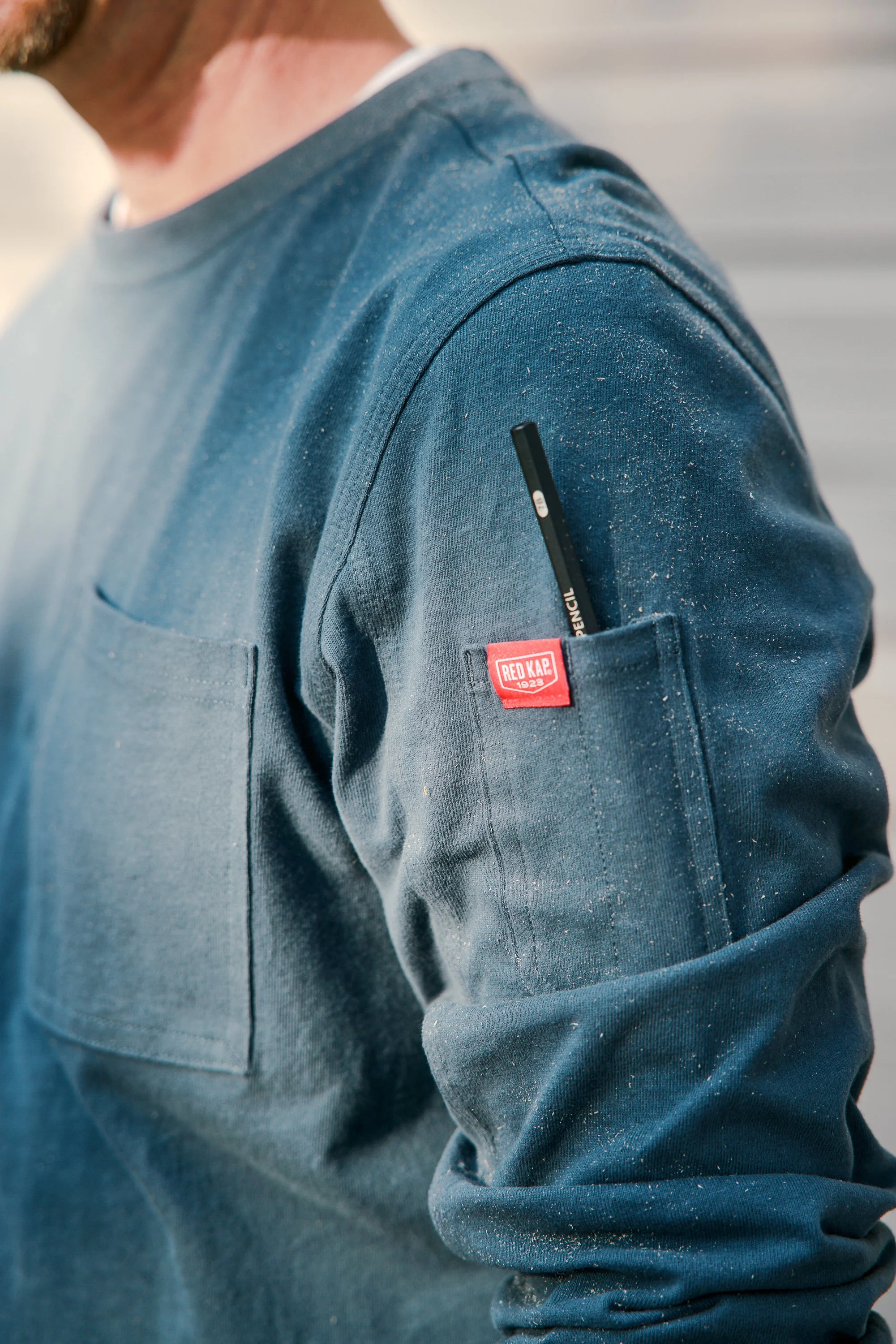 Close-up of a person wearing a blue jacket with a Red Kap tag, with a pencil in the jacket's pocket, possibly dusted with white powder or paint splatters.