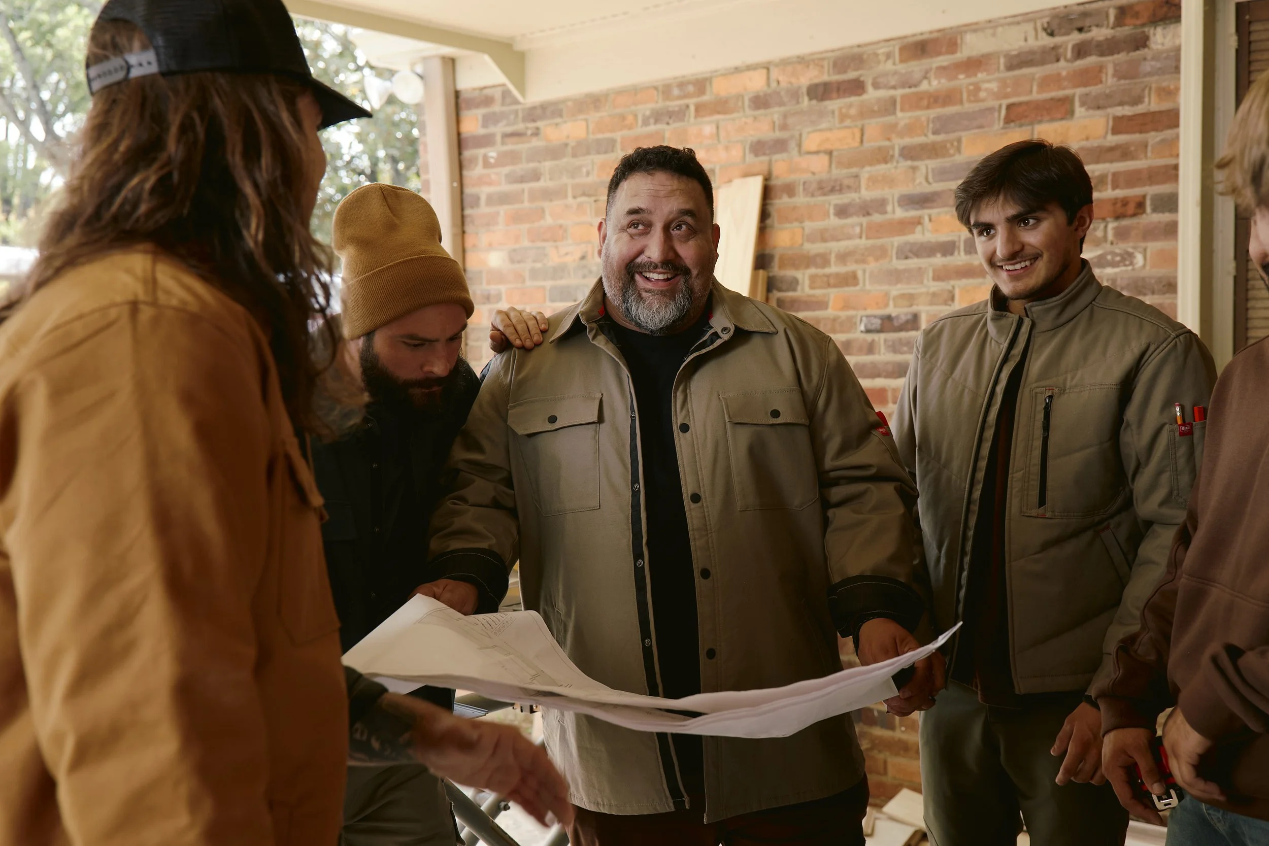A group of young men and an older man are gathered together outdoors, looking at blueprints or plans. They are smiling and appear to be having a discussion about construction or home improvement, with a brick wall and trees in the background.