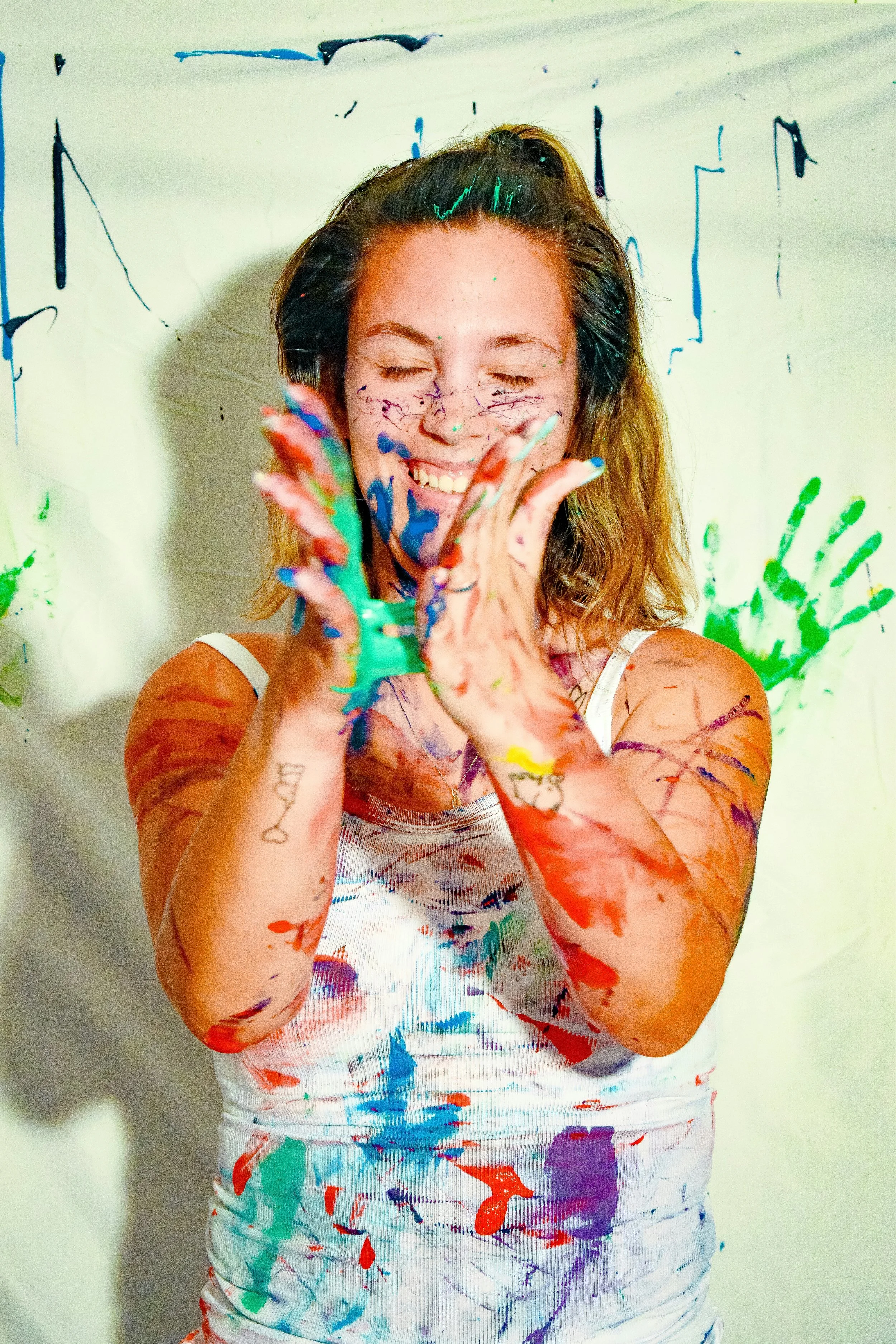 A woman with closed eyes, smiling, and covered in colorful paint splatters, standing against a white background also splattered with paint.