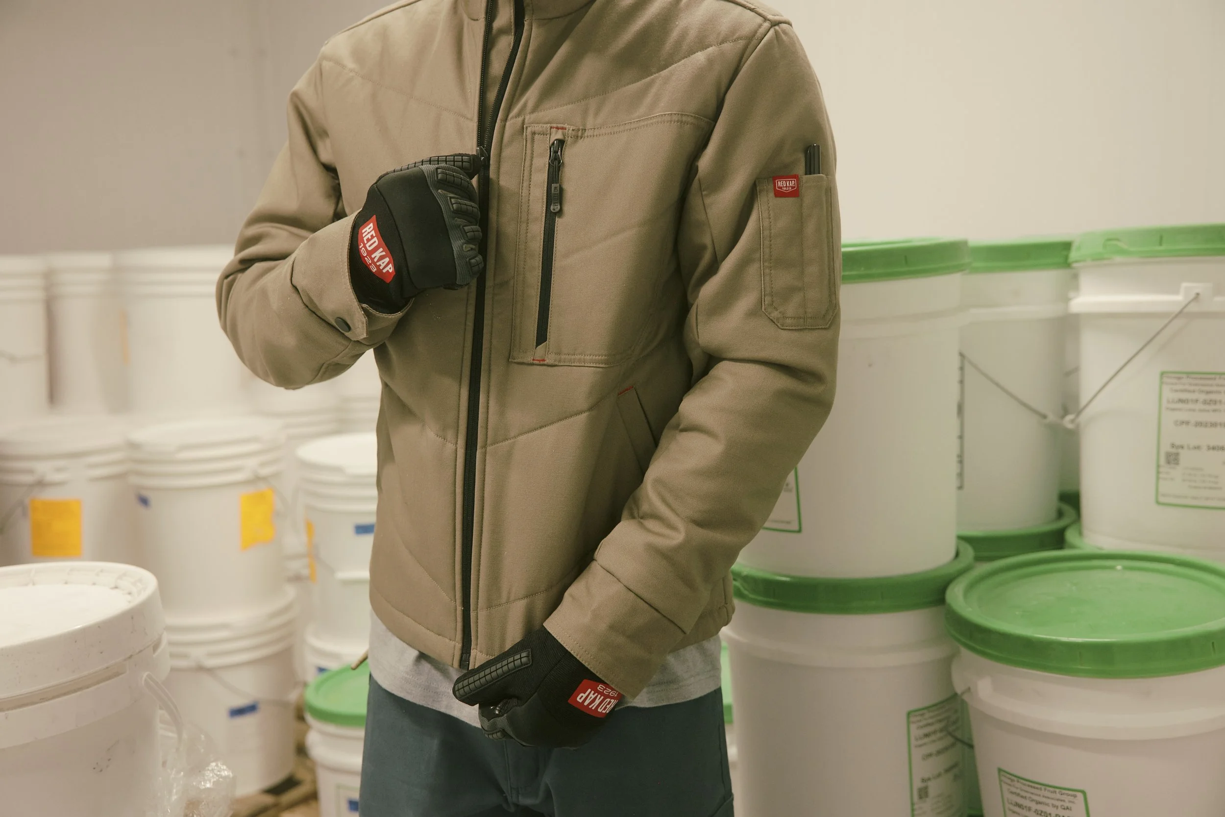 Person wearing a beige jacket and black gloves standing in front of white buckets with green-lidded buckets to the right. The person is holding their hand up close to their chest.