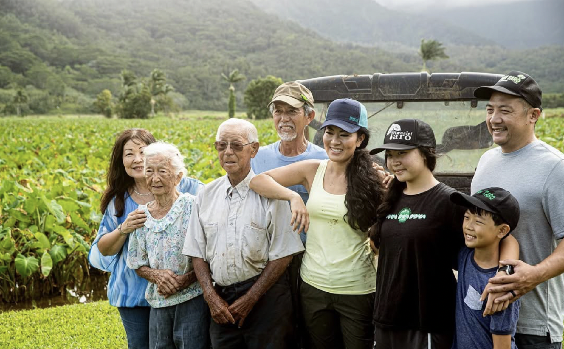 A group of diverse people, including elderly and children, smiling and standing together outdoors in a green field with mountains in the background during daytime.