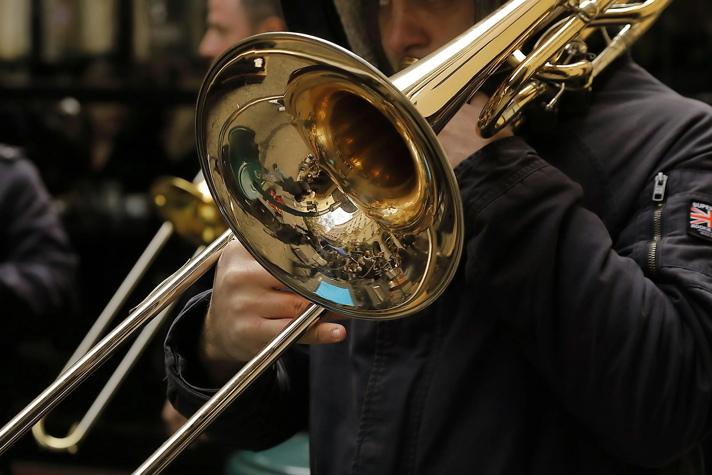 A person playing a shiny brass trombone during a street performance, reflected in the instrument's bell.