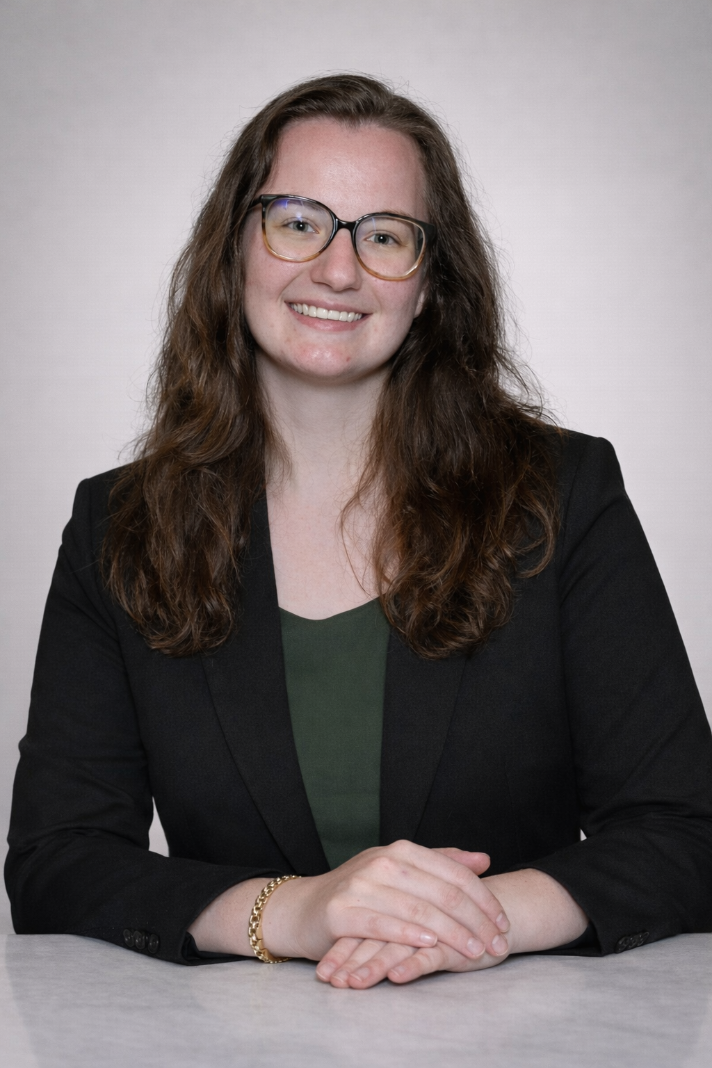 A woman with long curly brown hair, wearing glasses and a black blazer, sitting at a table with her hands folded, smiling at the camera.