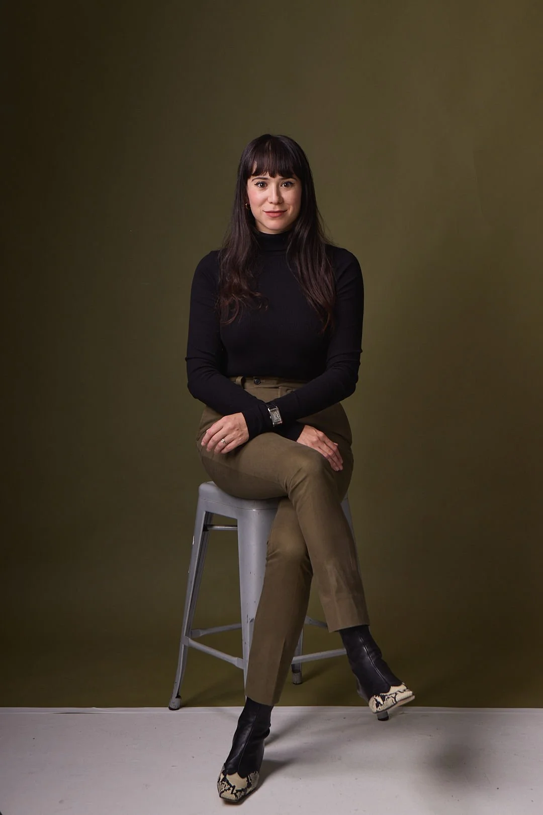 AJ Ayers, Writer, Speaker, and financial advisor, a  woman with long dark hair and bangs, wearing a black turtleneck sweater, khaki pants, and black and animal print boots, sitting on a stool against an olive green background.
