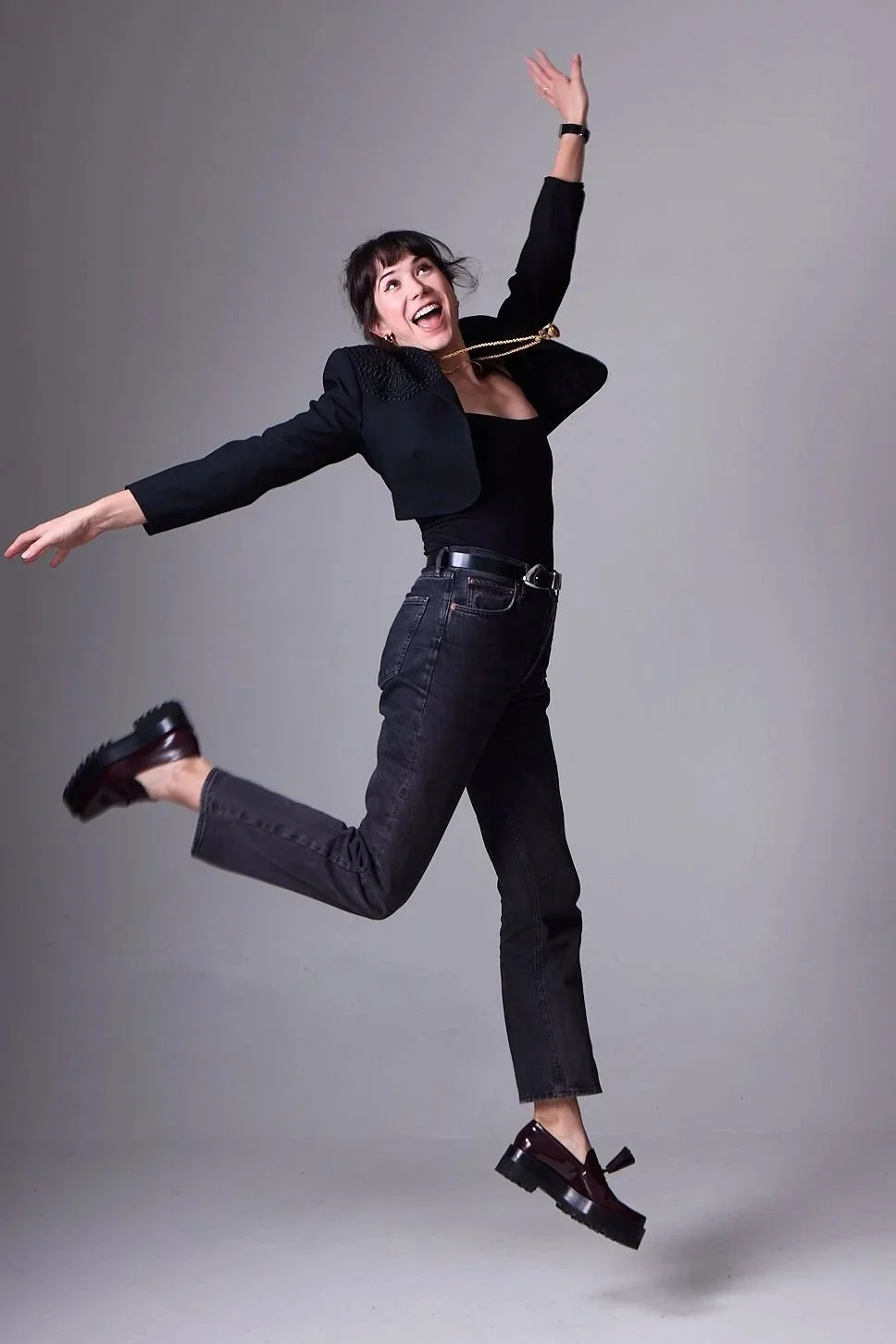 AJ Ayers, Writer, Speaker, and financial advisor, a woman in black clothing joyfully jumping with arms outstretched against a gray background.
