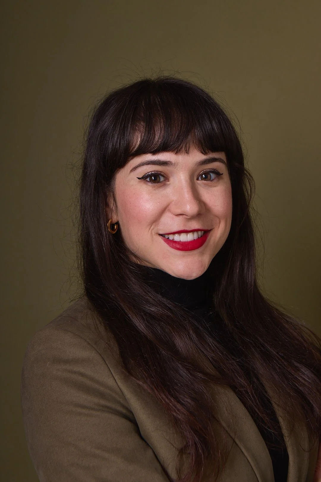 AJ Ayers, Writer, Speaker, and financial advisor, a woman with dark brown hair and bangs, wearing a black turtleneck and brown blazer, smiling at the camera, against a plain olive-green background.