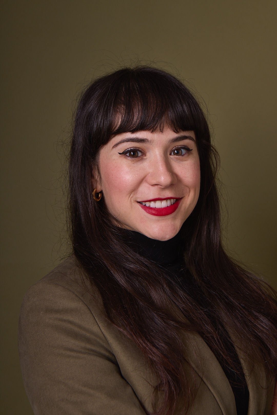 AJ Ayers, Writer, Speaker, and financial advisor, a woman with long brown hair, wearing a black turtleneck and khaki blazer, smiling at the camera, against a plain olive green background.