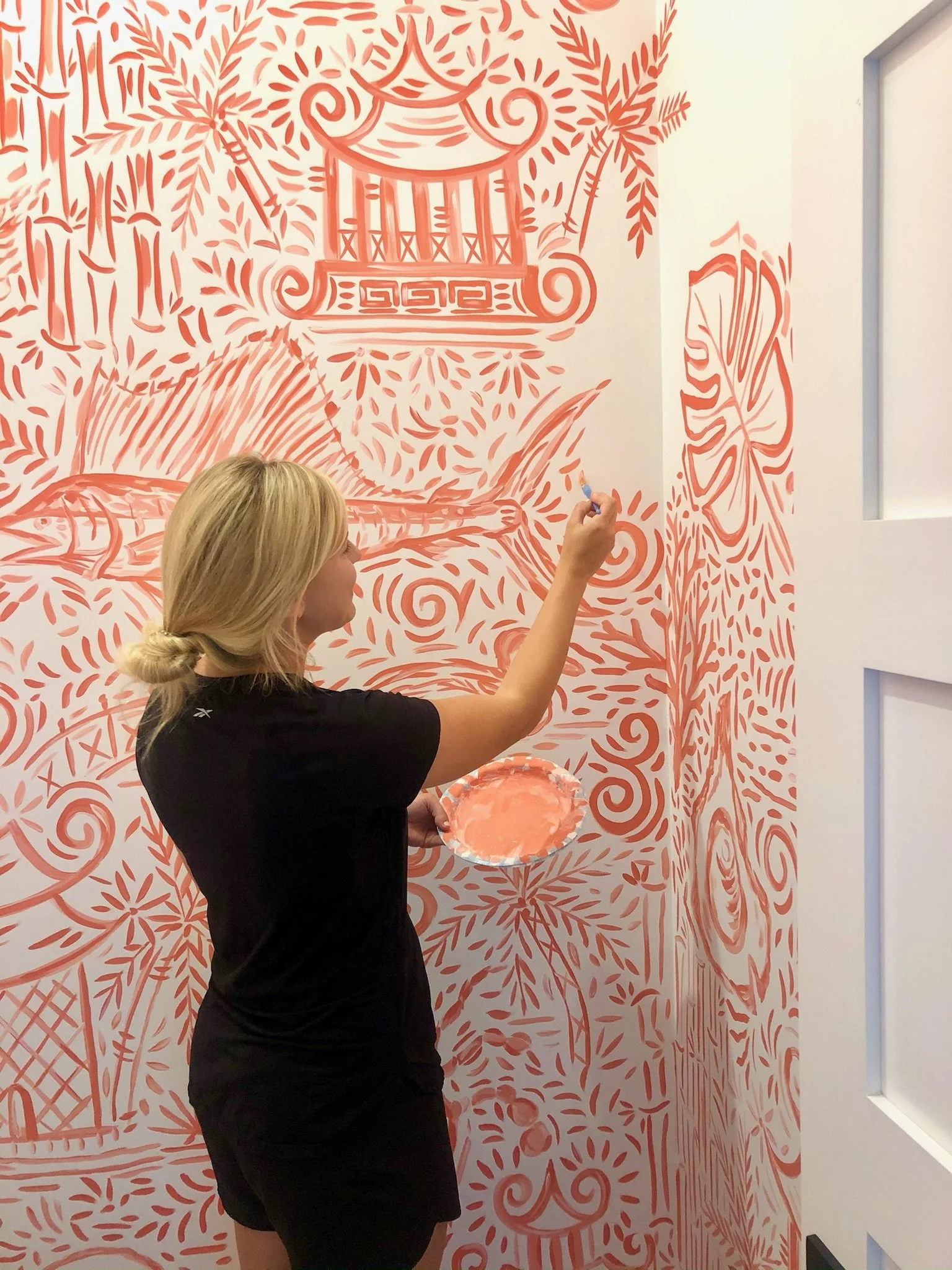 A woman is painting a mural with a peach color on a white wall, featuring tropical elements such as a pagoda, fish, and plants.