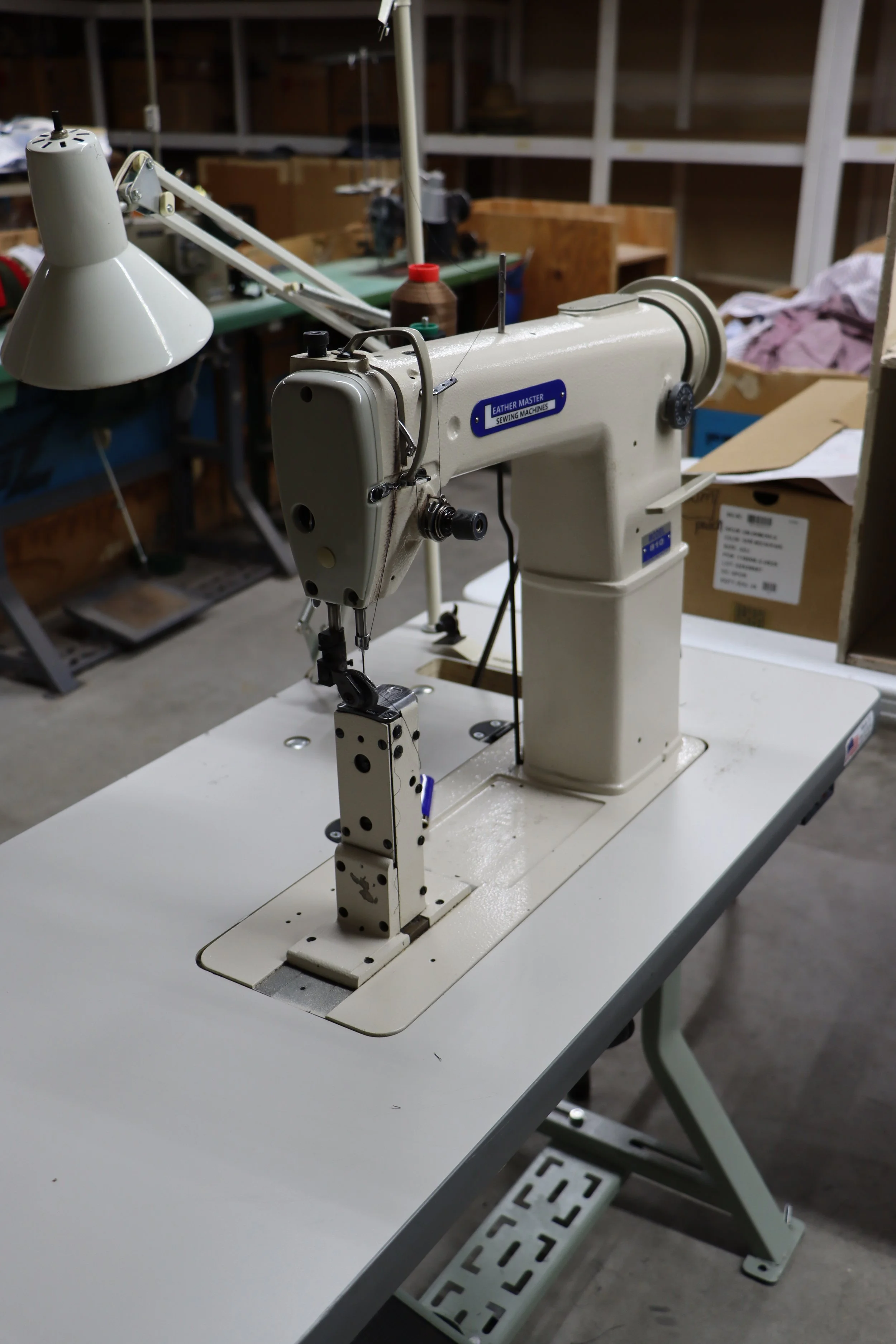 A vintage industrial sewing machine with a white body on a white table in a workshop, with shelving and supplies in the background.
