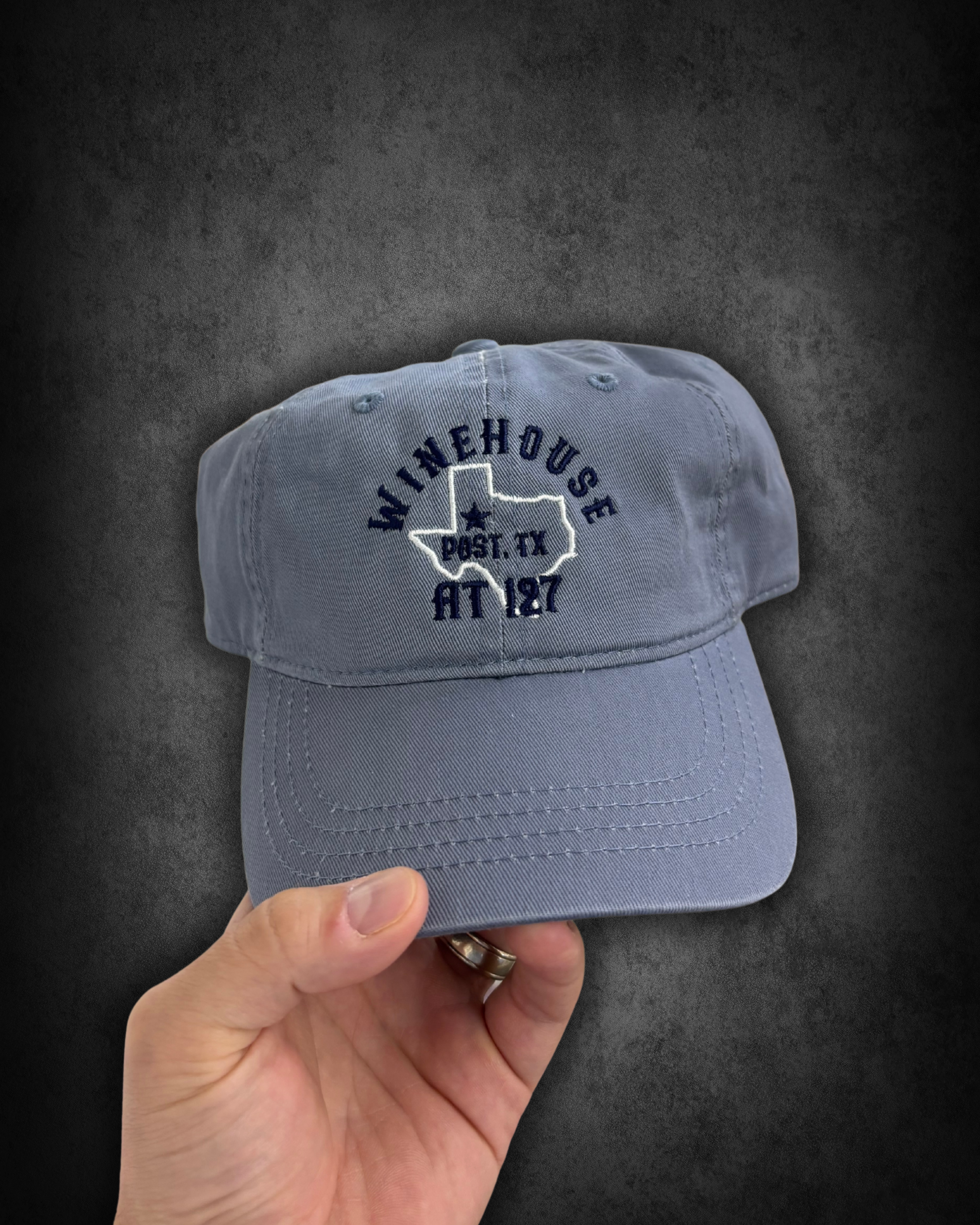 Gray baseball cap with embroidery of the state of Texas and the words 'WINEHOUST' and 'PAST. TX' on the front, held by a person's hand against a dark background.