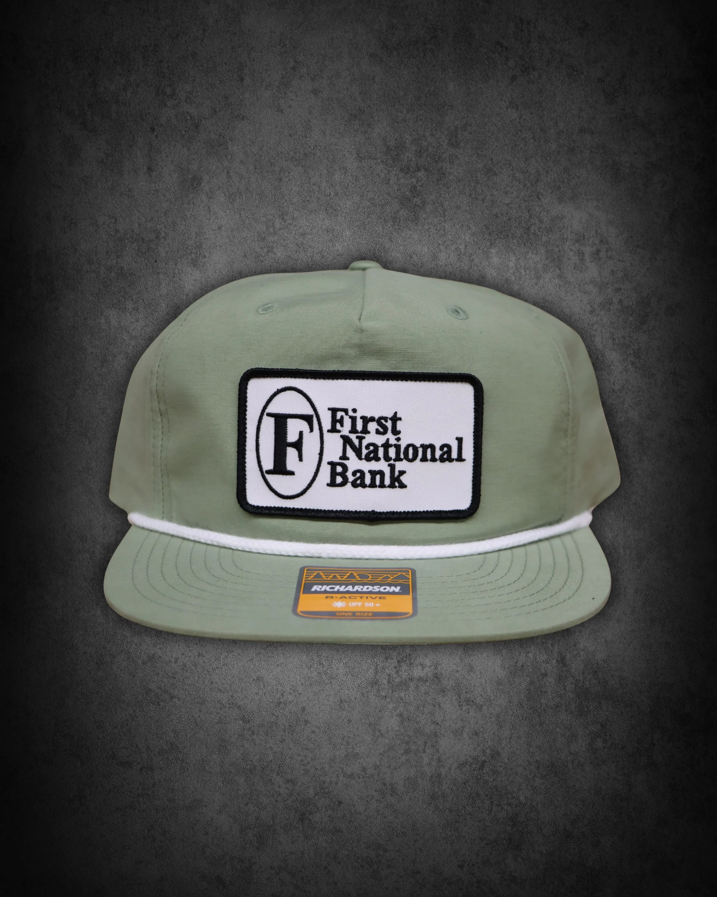 Front view of a beige Richardsons RACTIVO cap with a black and white First National Bank patch on the front, placed on a black surface.