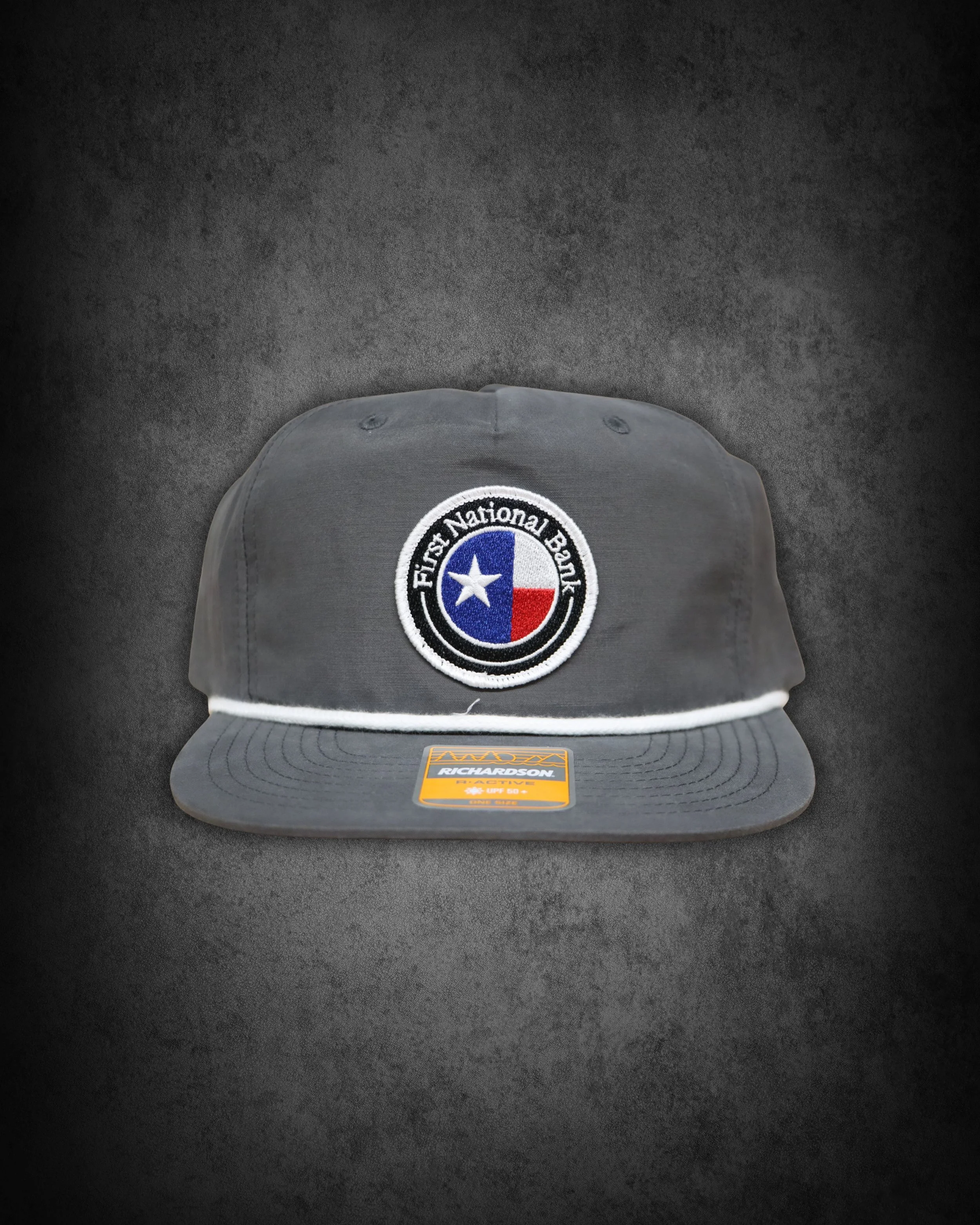 Gray baseball cap with a Texas flag emblem and the words "First National Bank" on the front, placed on a dark surface.