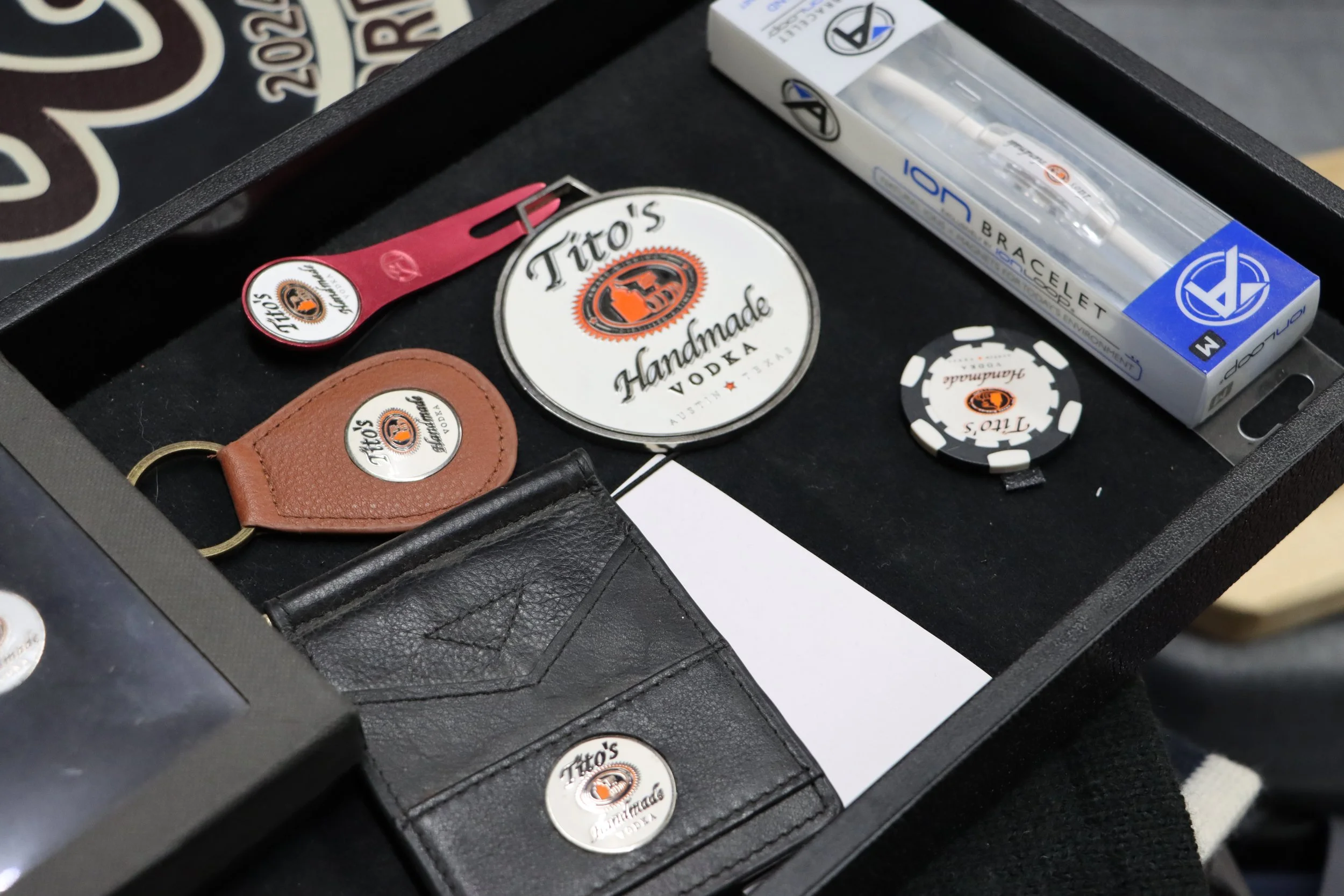 A black display case holding Tito's Handmade Vodka accessories and promo items, including a coaster, keychain, and more Tito's vodka promo items.