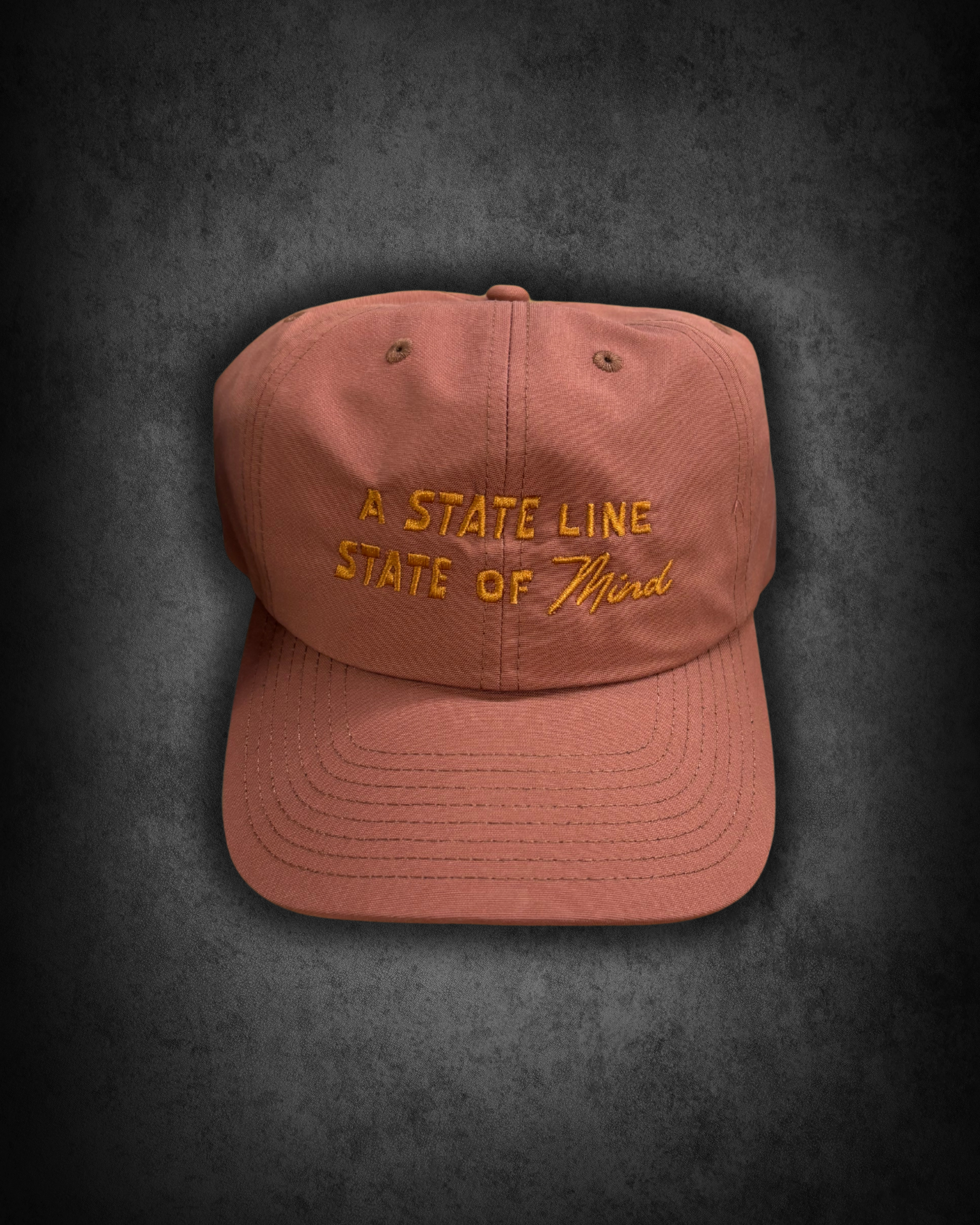 Pink baseball cap with embroidered text that reads "A STATE LINE STATE OF Mind," placed on a dark surface.