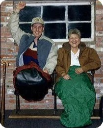 Two young people sitting on a bench indoors, smiling, with a brick wall and window in the background. The person on the left is waving and wearing a cap and jacket, holding a backpack. The person on the right is wearing a green dress or skirt with a cardigan.