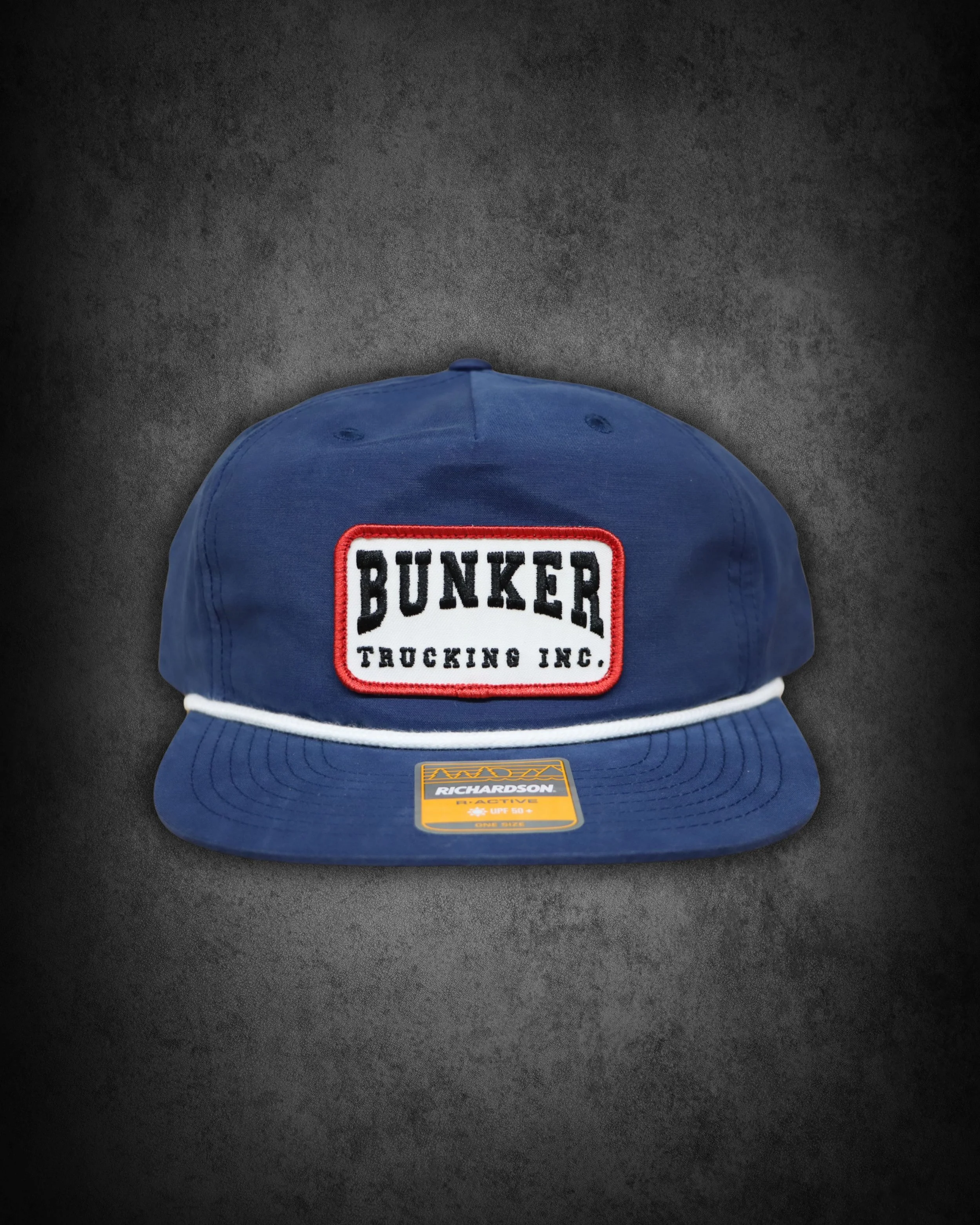 Blue trucker cap with patches, one says 'BUNKER TRUCKING INC.' and another indicates 'Richardson' brand with 'UPF 50' and 'ONE SIZE' details, placed on a dark surface.