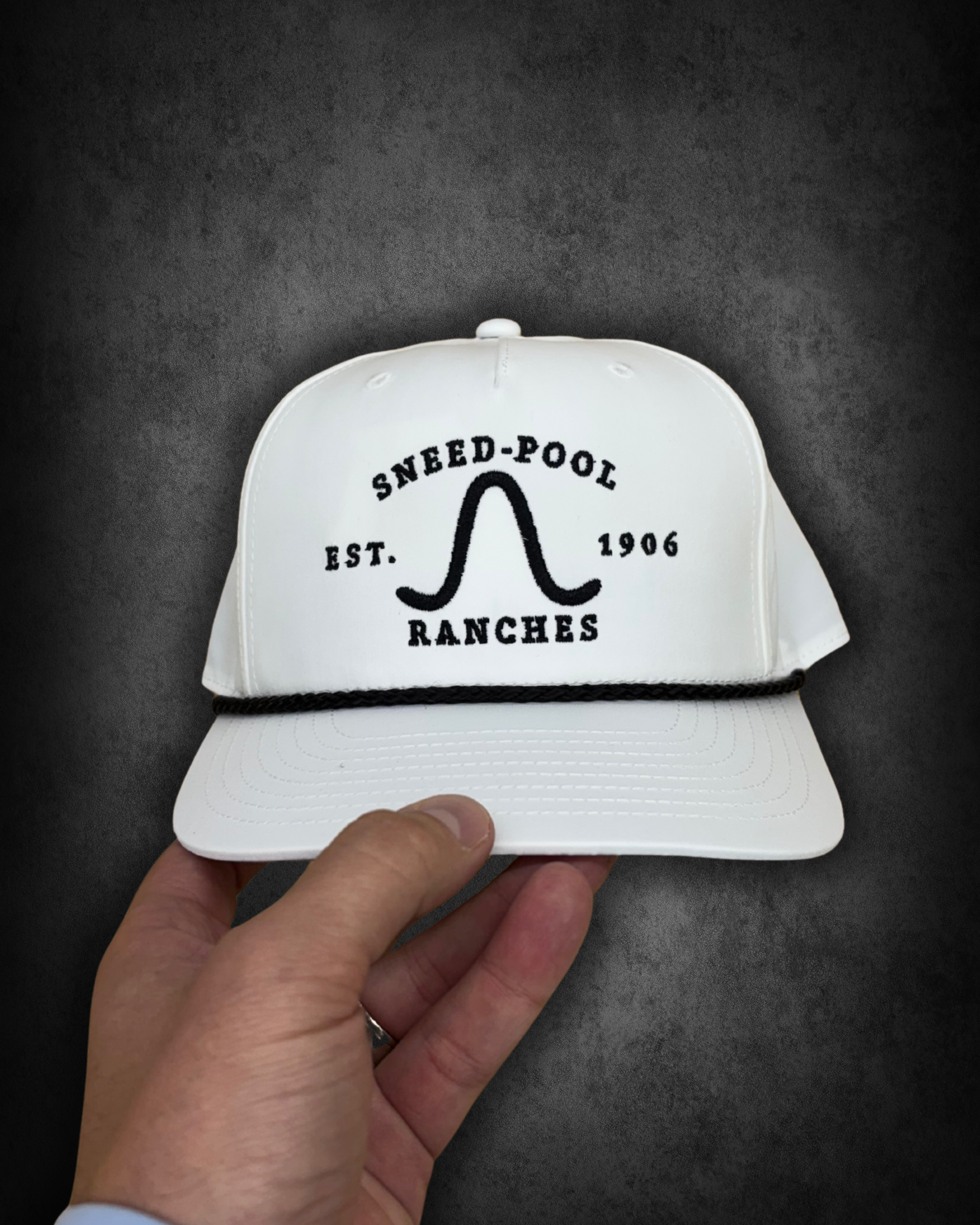 A person holding a white trucker cap with black embroidered text that reads 'SNEED-POOL EST. 1906 DANCES RANCHES' against a dark background.