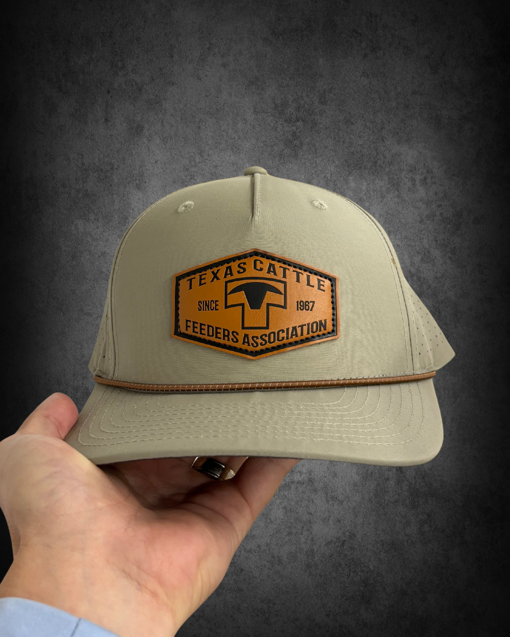 A beige baseball cap with a patch reading 'TEXAS CATTLE FEEDERS ASSOCIATION' worn by a hand against a dark gray background.