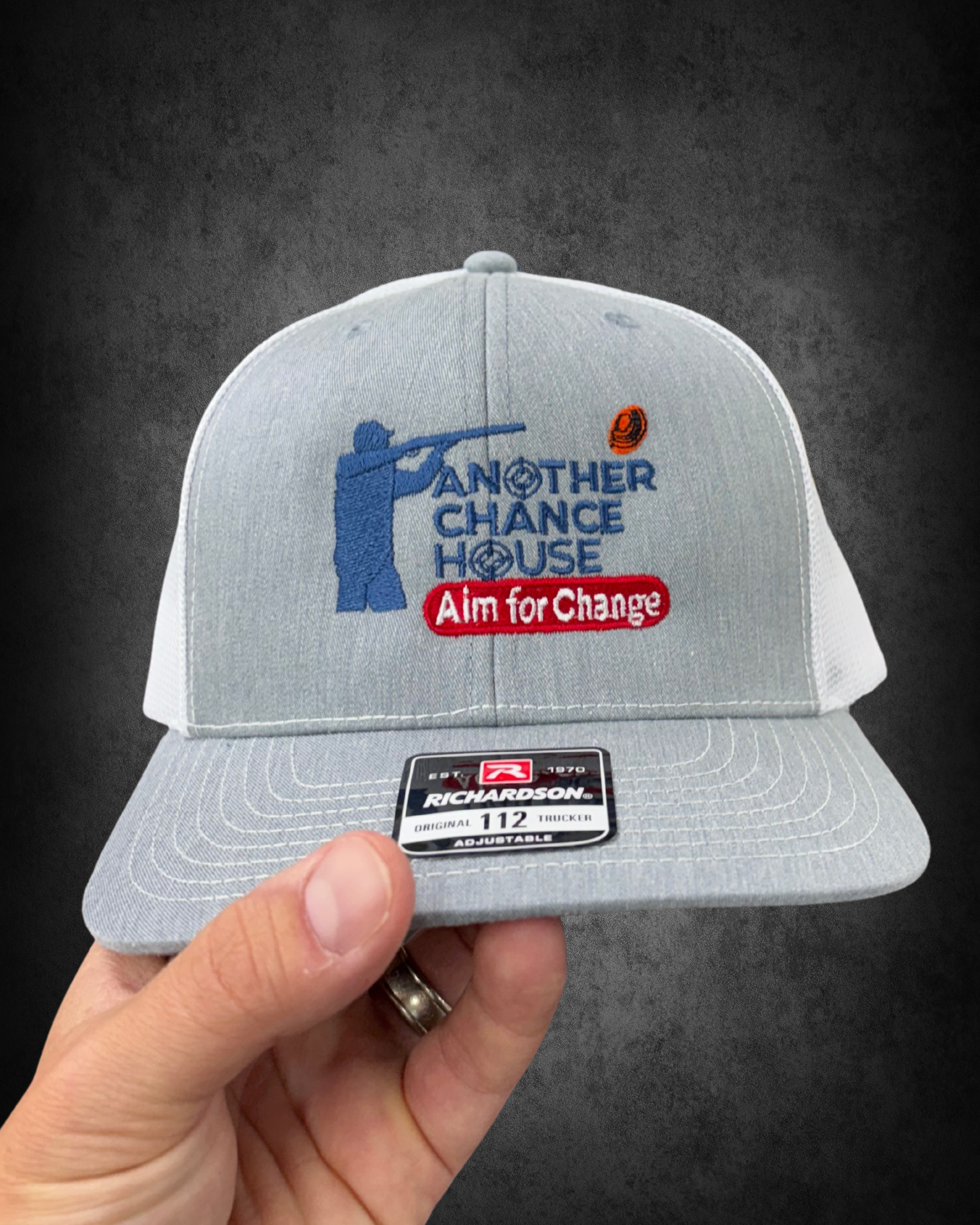 Gray trucker hat with embroidered phrase 'Aim for Change' above a baseball and the words 'Another House' on the front. The hat has an adjustable strap and is being held by a person's hand against a dark surface background.