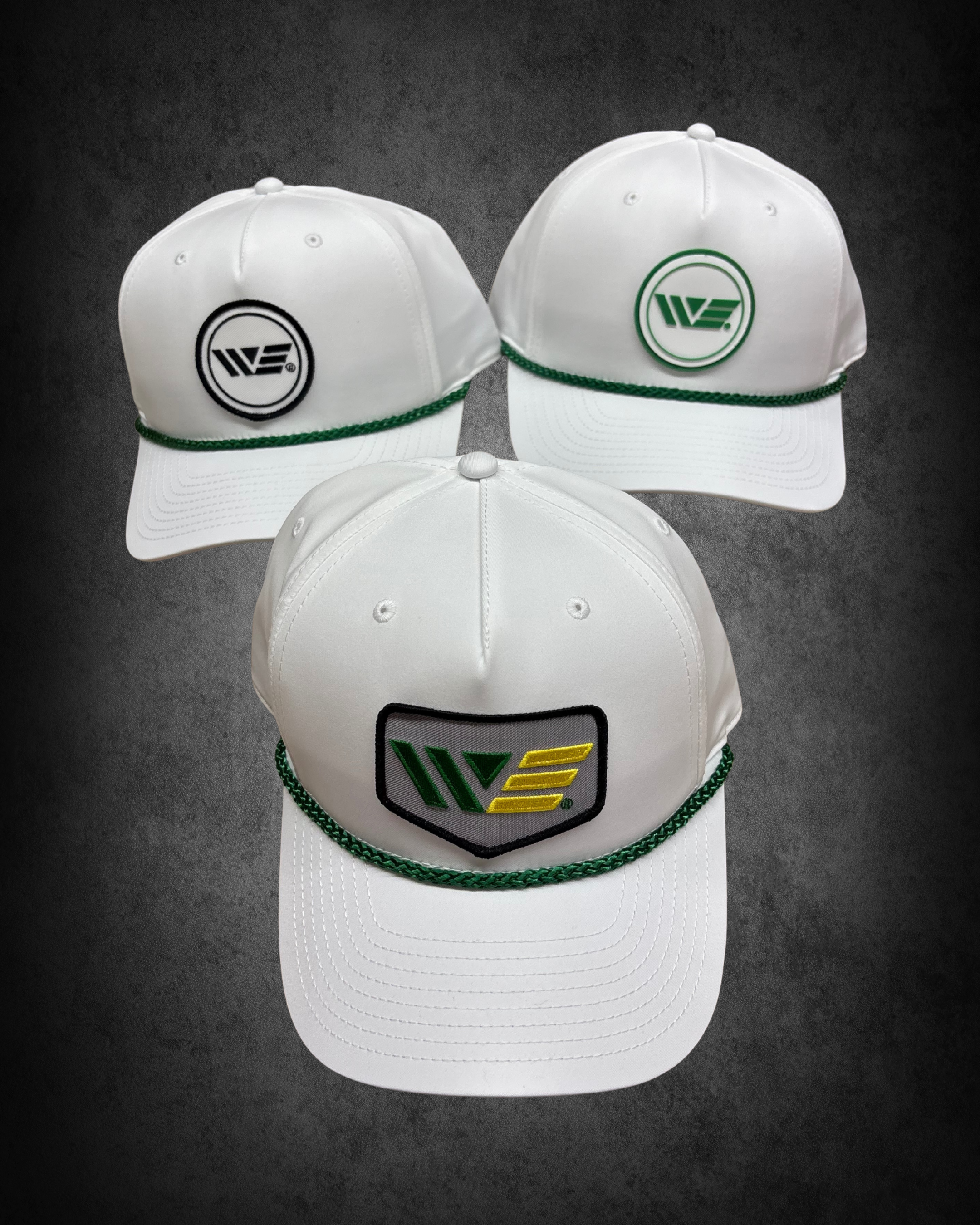 Three white baseball caps with green and black logos on the front, arranged on a dark background. The caps have a green braided trim around the base and are visible from a top-front angle.