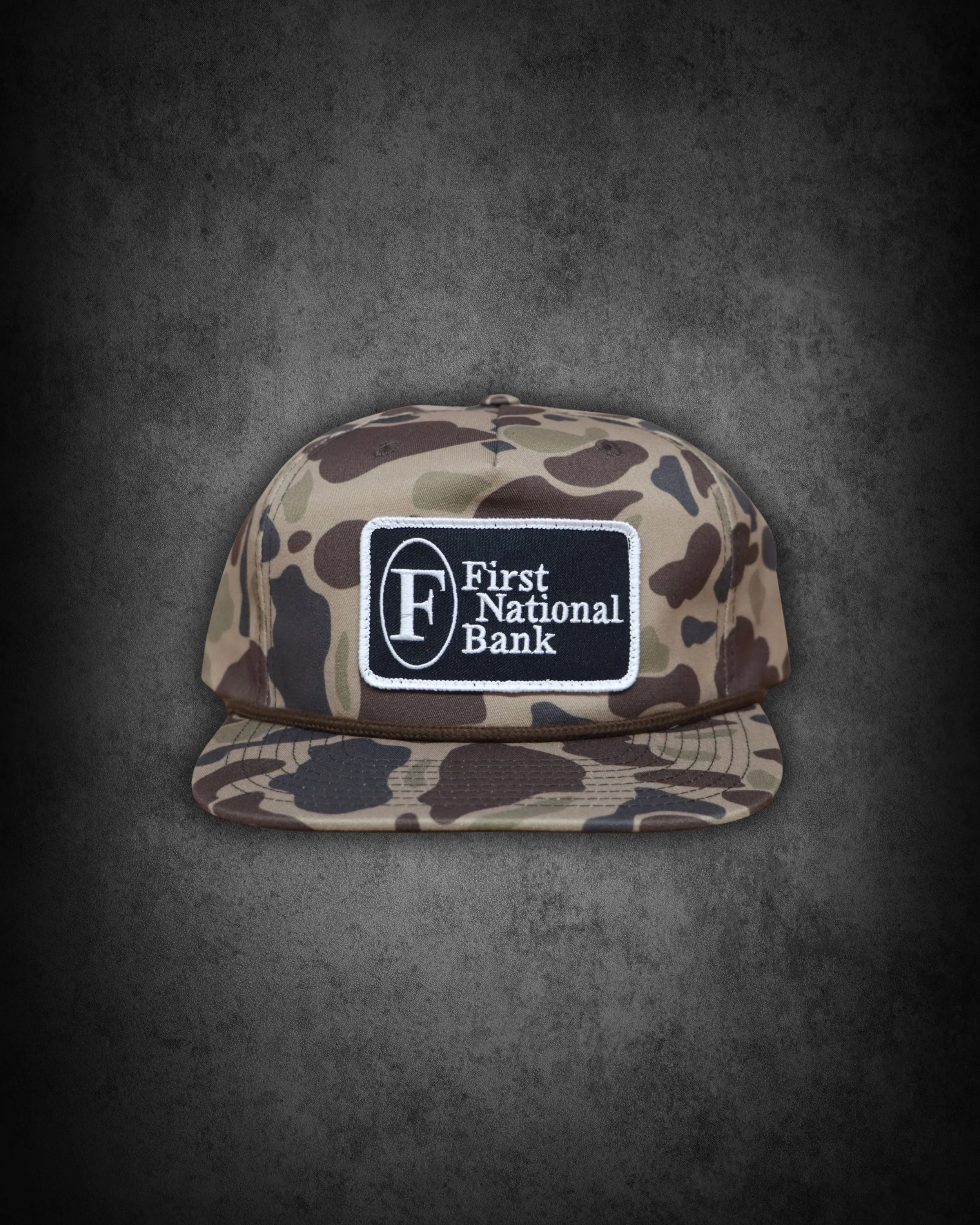 A camouflage patterned baseball cap with a rectangular patch on the front that has the logo and name of First National Bank.