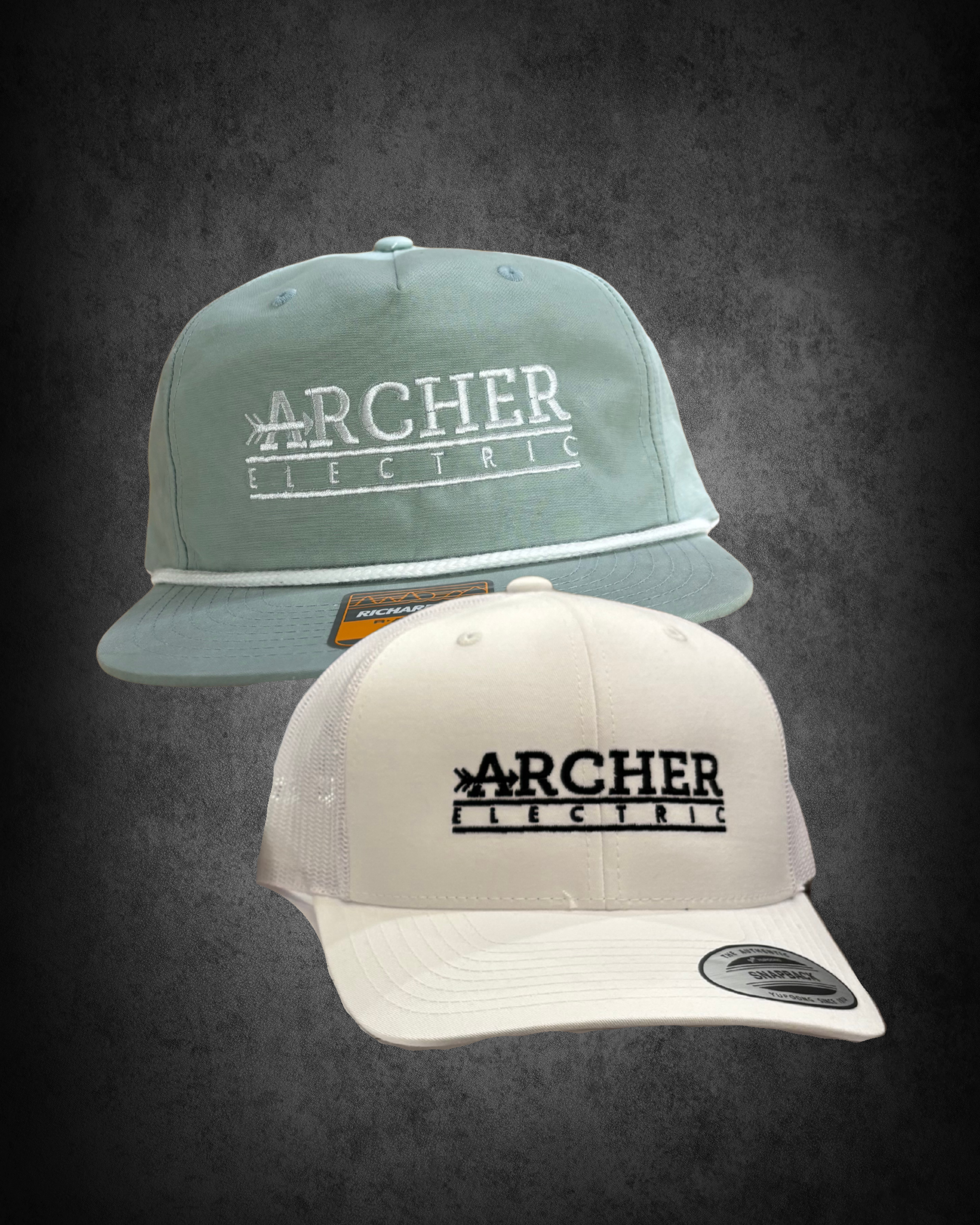Three baseball caps stacked one behind another on a dark surface. The top cap is light blue with 'ARCHER ELECTRIC' embroidery, the middle cap is light gray also with 'ARCHER ELECTRIC' embroidery, and the bottom cap is white with the same embroidery.