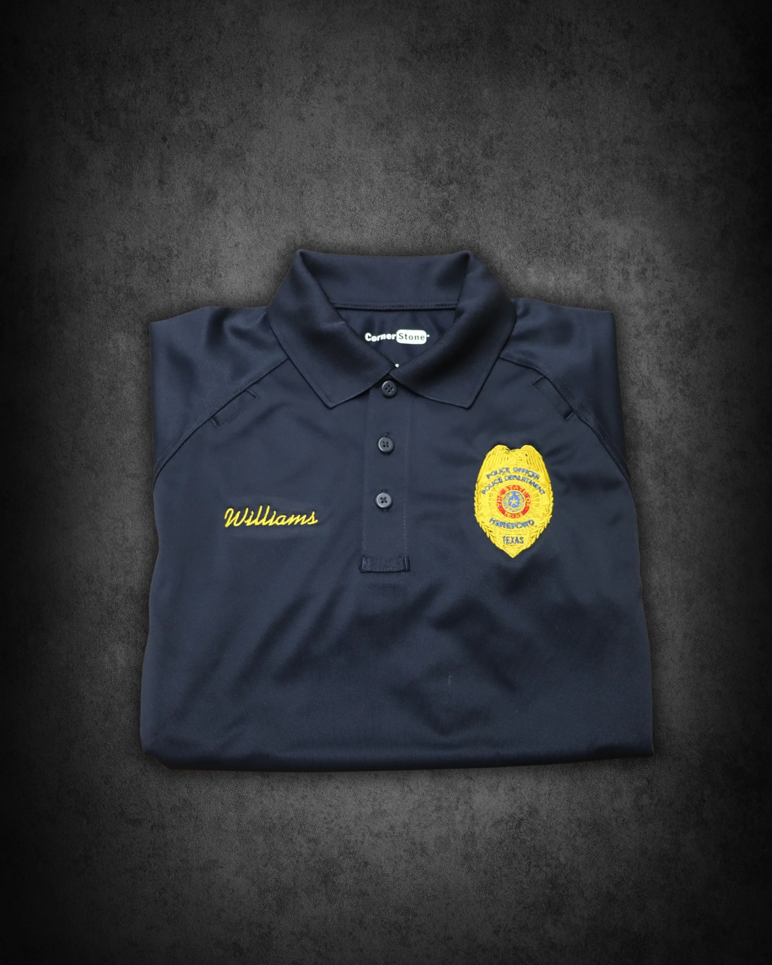 A folded navy blue police uniform shirt with a badge and embroidered name 'Williams' on the front, placed on a dark surface.