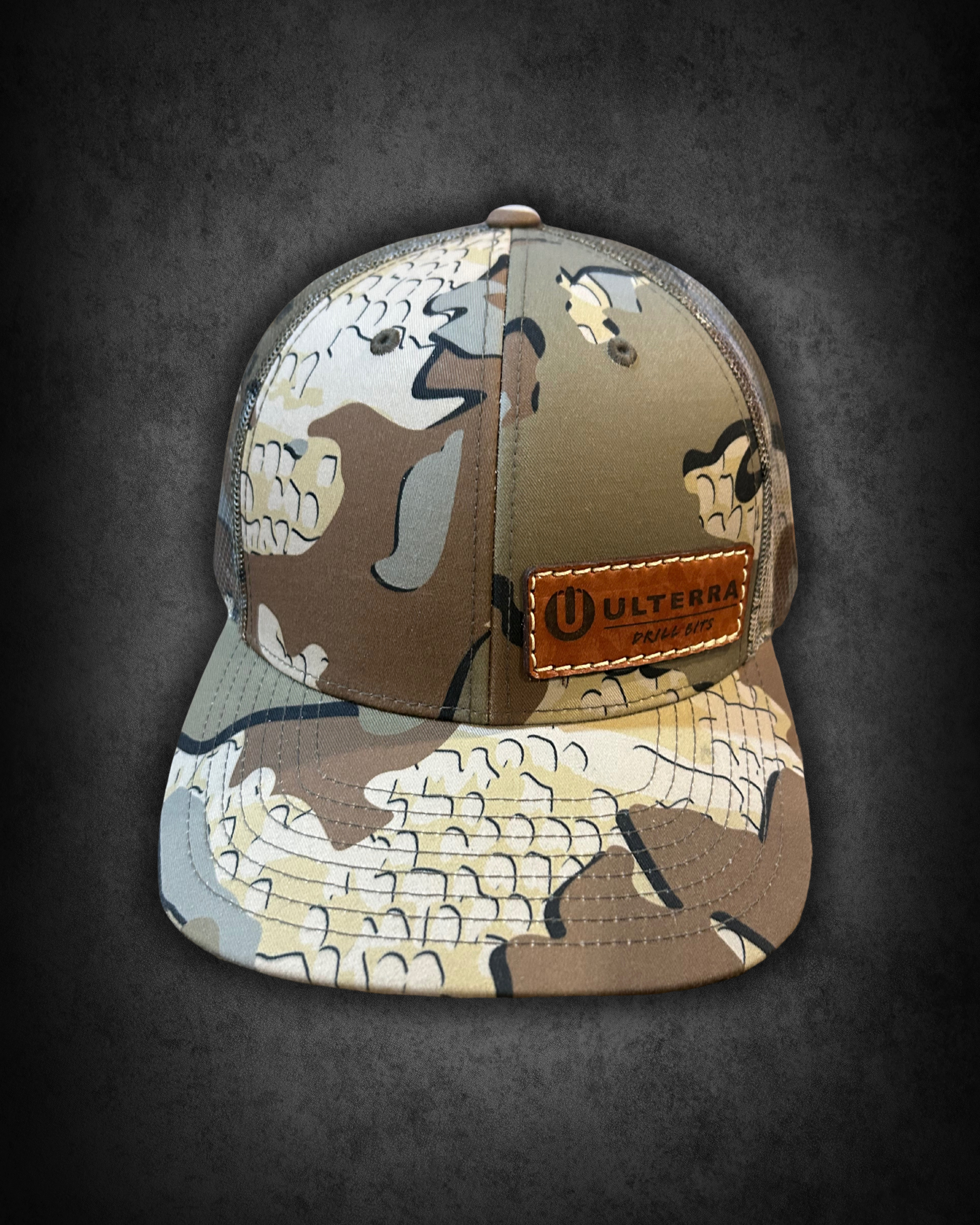 Camouflage baseball cap with a leather patch that reads "ULTERRA" on the front, placed on a dark surface.