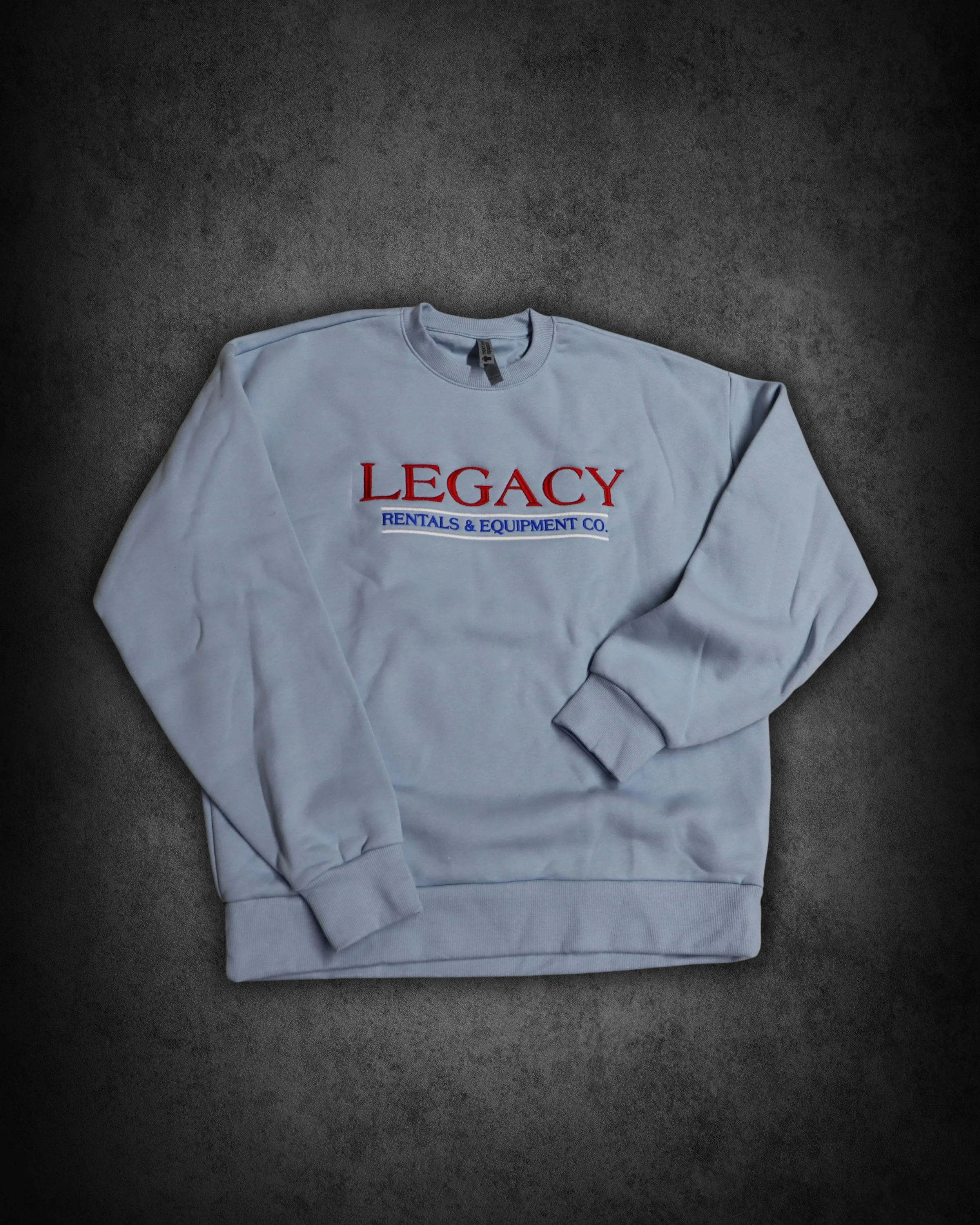 A light gray sweatshirt with the text 'LEGACY RENTALS & EQUIPMENT CO.' printed on it in red and blue letters, lying on a dark surface.
