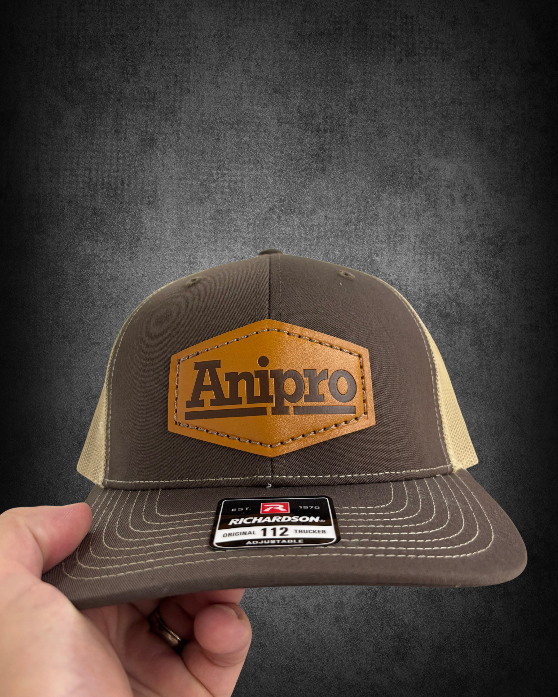 Close-up of a baseball cap with a brown leather patch that reads "Antiro" on the front. The cap is brown with beige sides and has a sticker that indicates it's a Richardson brand trucker hat, size 112, adjustable. The background is a dark gray or bla