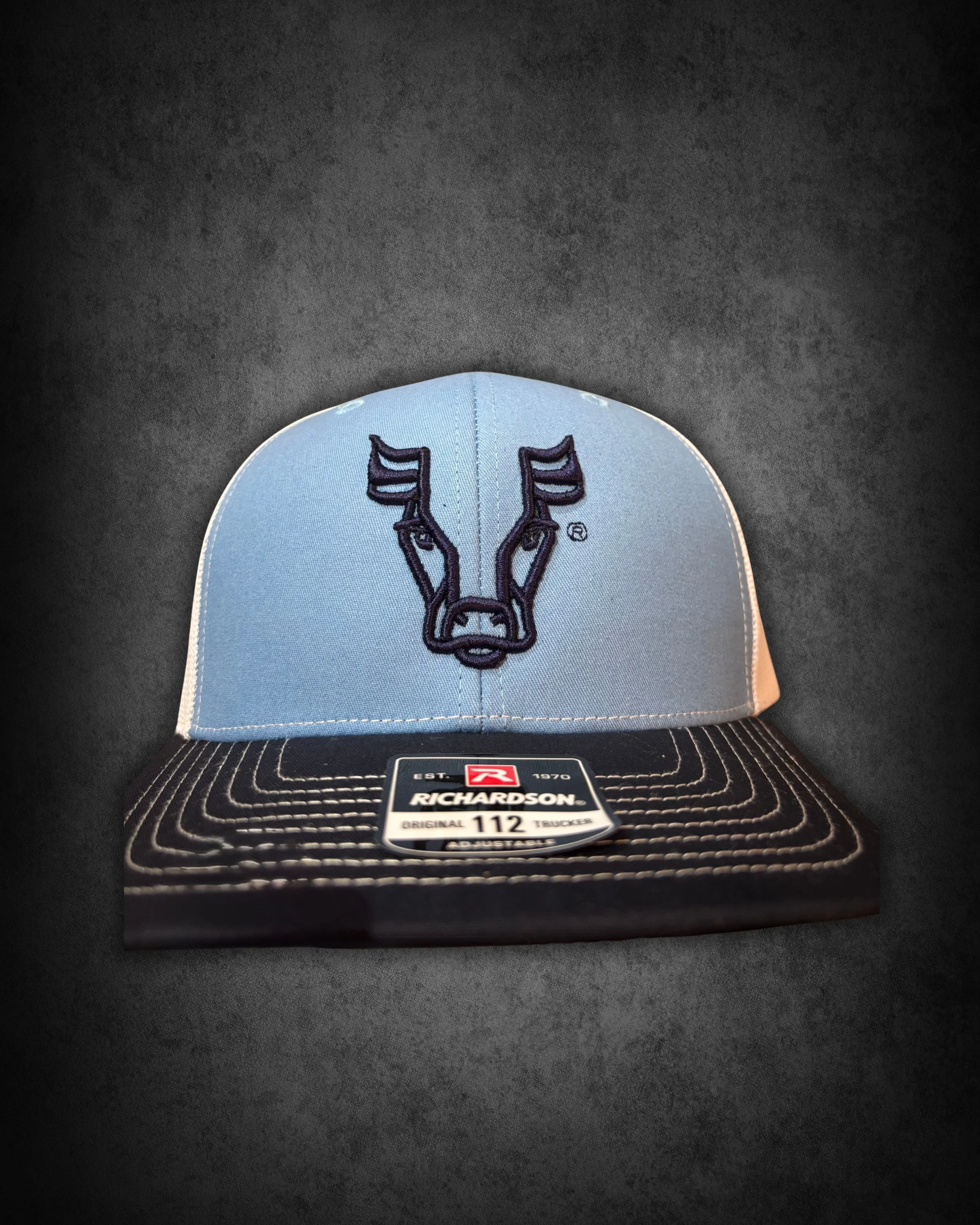 A blue Richardson trucker cap with black and white stitching and an embroidered deer head logo on the front, positioned on top of a black textured surface store tag that says Richardson and size 112.