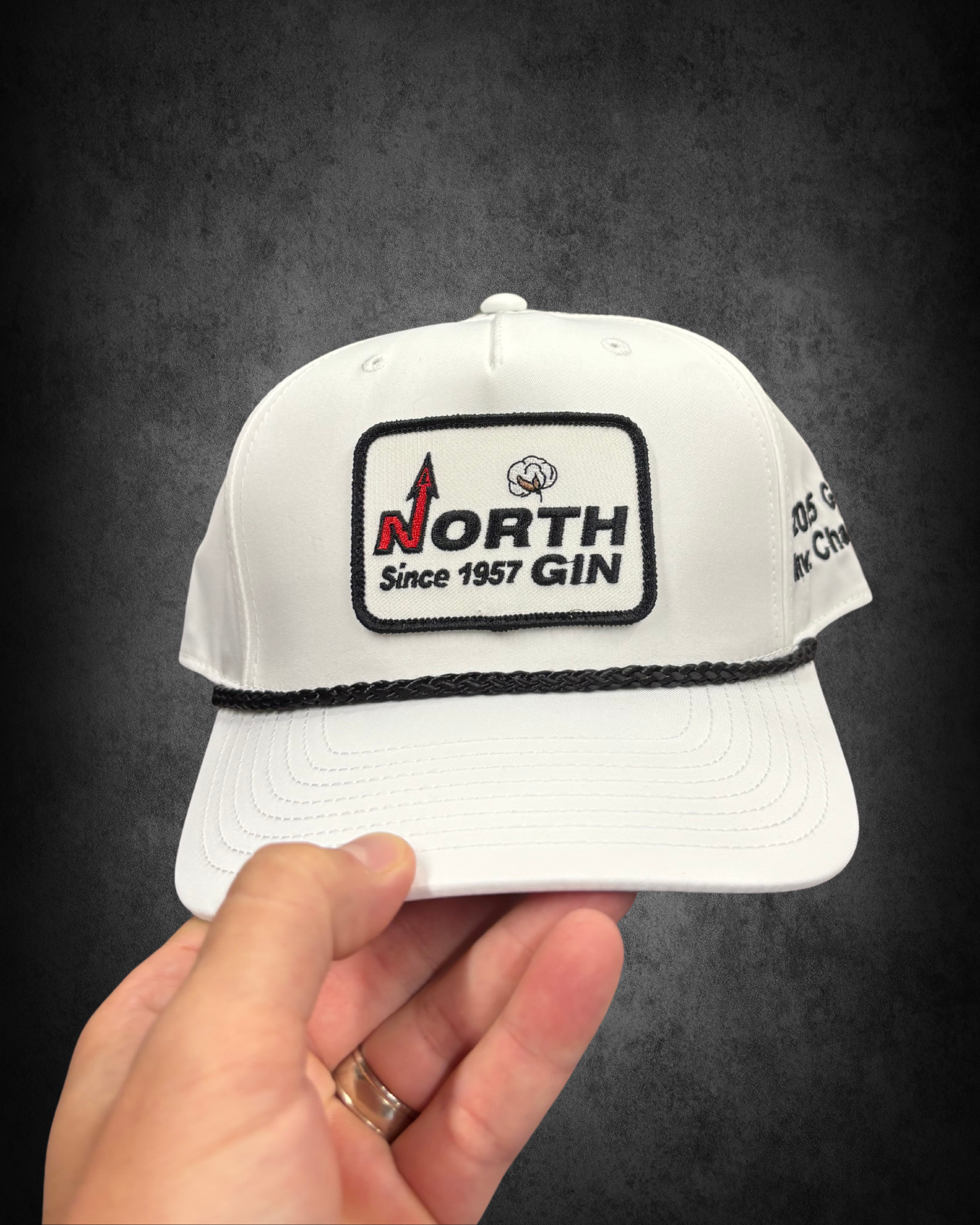 Hand holding a white baseball cap with a logo that reads 'North Since 1957 Gin' and features a cloud and a red upward arrow, against a black background.