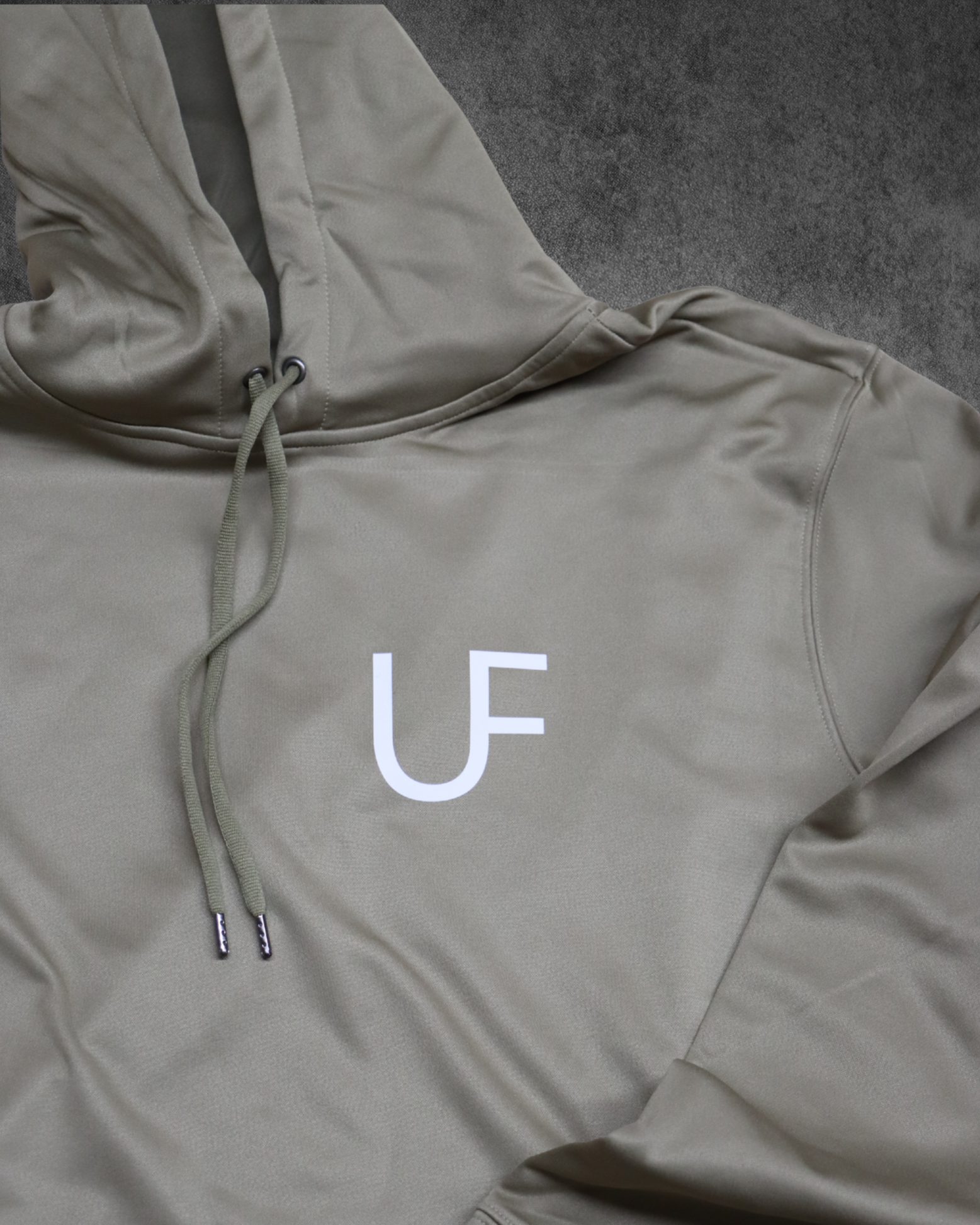 Close-up of a beige hoodie with a partial white logo, drawstrings hanging down, and a dark gray background.