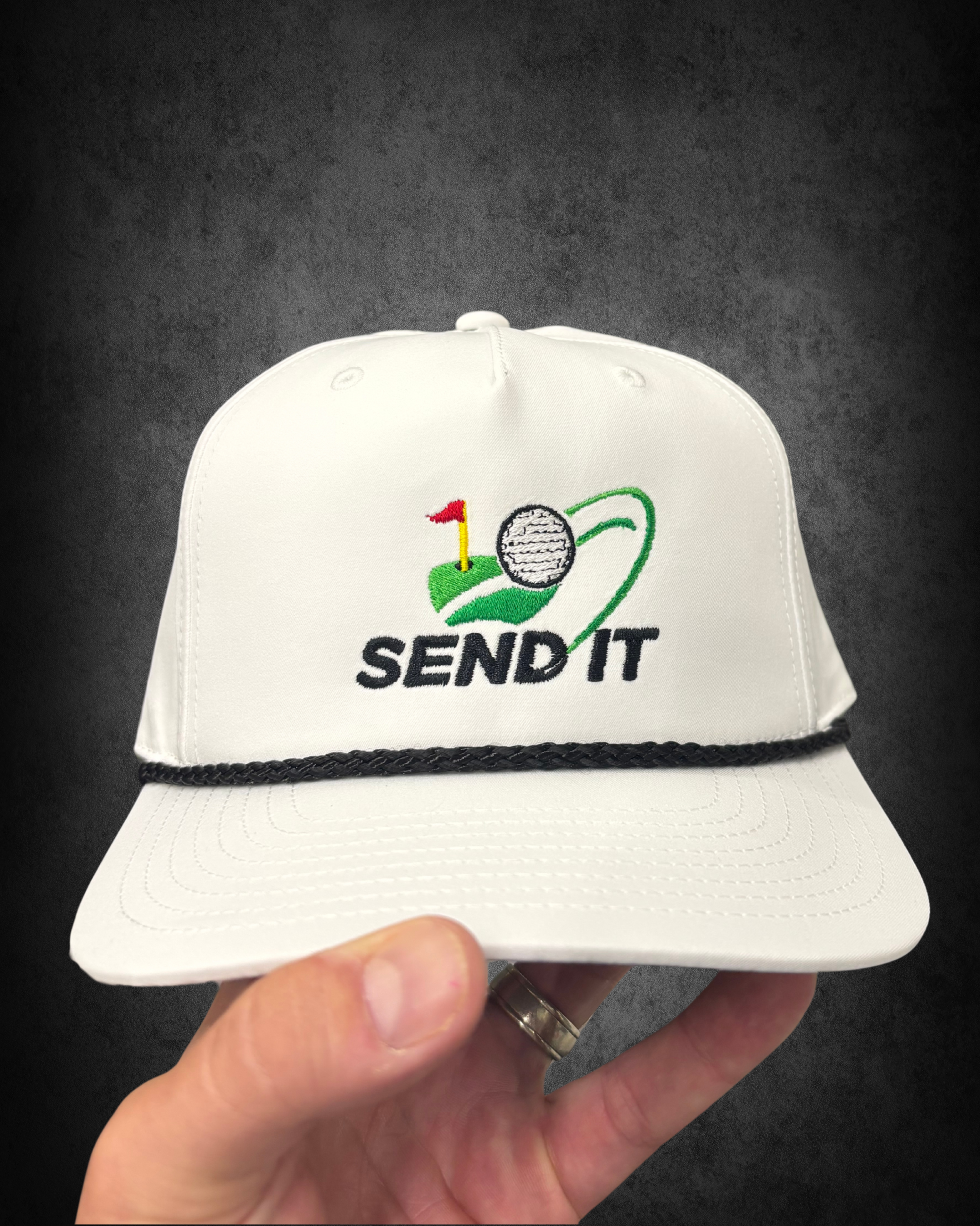 A white baseball cap with embroidered design featuring a golf ball, flag, and golf club, and the text 'SEND IT'. The cap has front panel stitching, eyelets, and a black braided strap on the rim.
