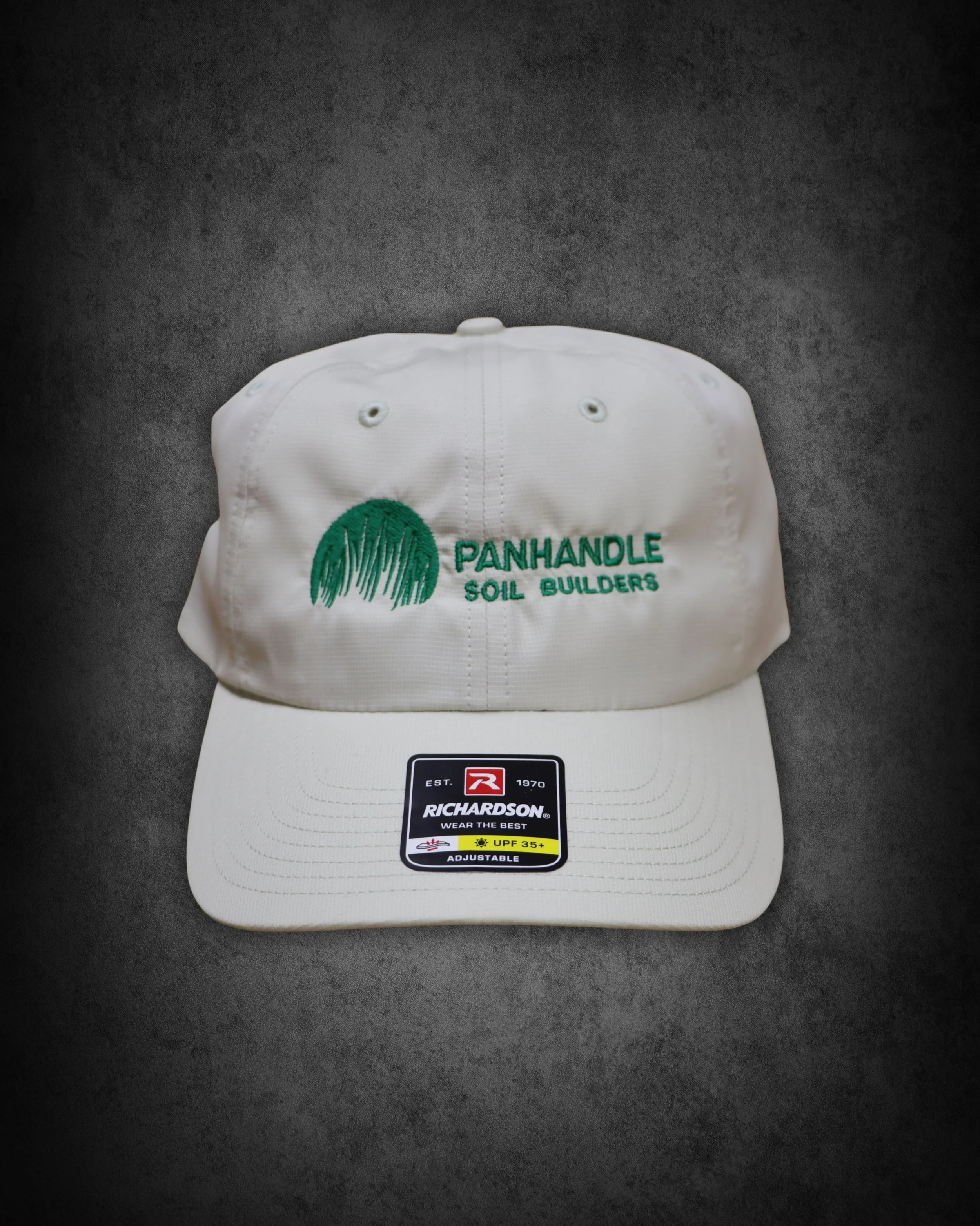 A white Richardson adjustable baseball cap with green embroidery featuring the words 'Panhandle Soil Builders' and an image of a soil mound with grass. The cap is placed on a white platform on a dark, textured surface.