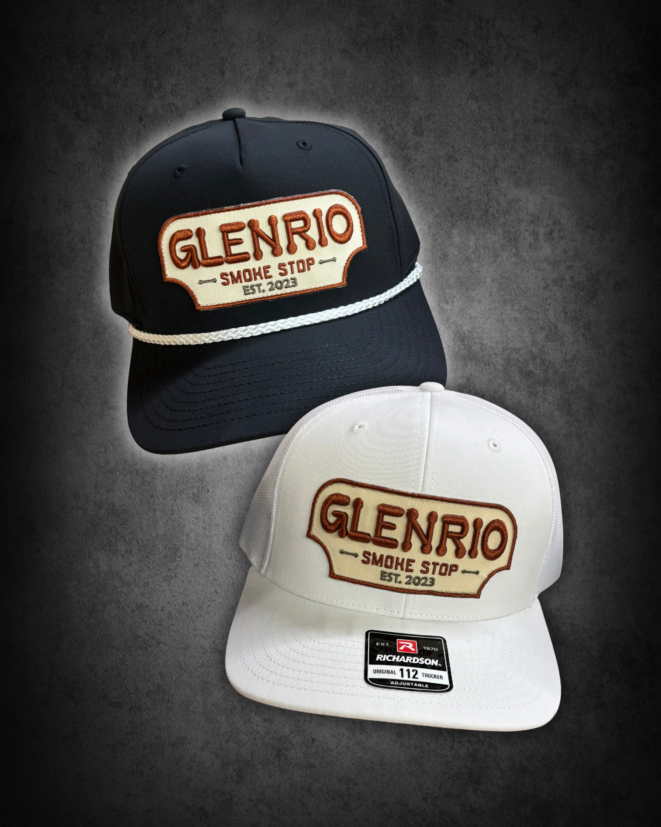 Three baseball caps with embroidered patches on the front, featuring the words "Glenrio Smoke Stop Est. 2023." The caps are black and white, with the black one placed in the back and the white one in the front, on a dark textured background. The whit
