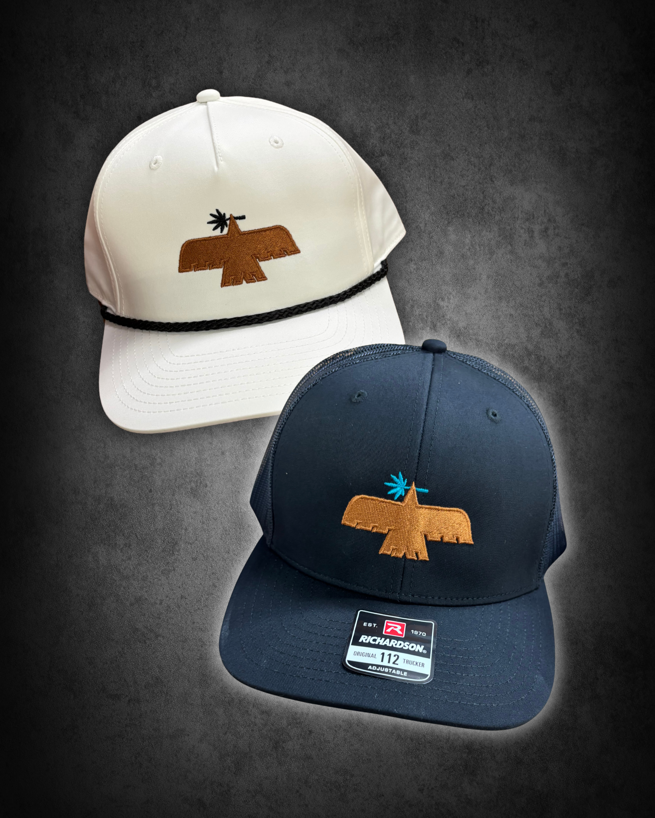 A white baseball cap with an embroidered brown eagle logo on the front and a black braided strap around the brim, placed on a dark surface. Partially visible navy blue cap with a similar logo is also in the image, with a Richardson brand tag on the b
