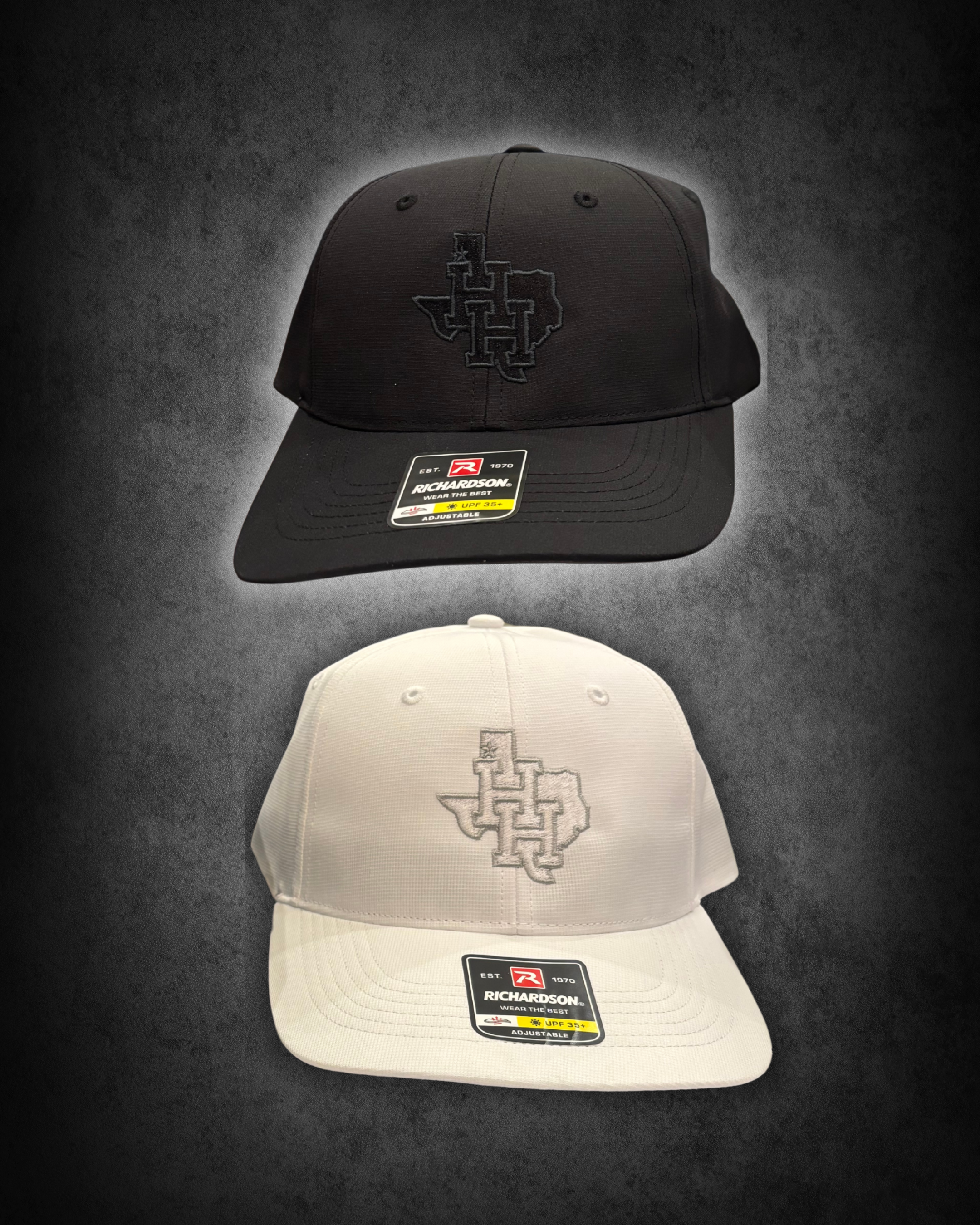 Two baseball caps, one black and one white, laid on a dark surface with a gray background. The black cap has a logo embroidered on the front and a sticker on the brim indicating it is adjustable and UPF 35+. The white cap has an embroidered logo on t