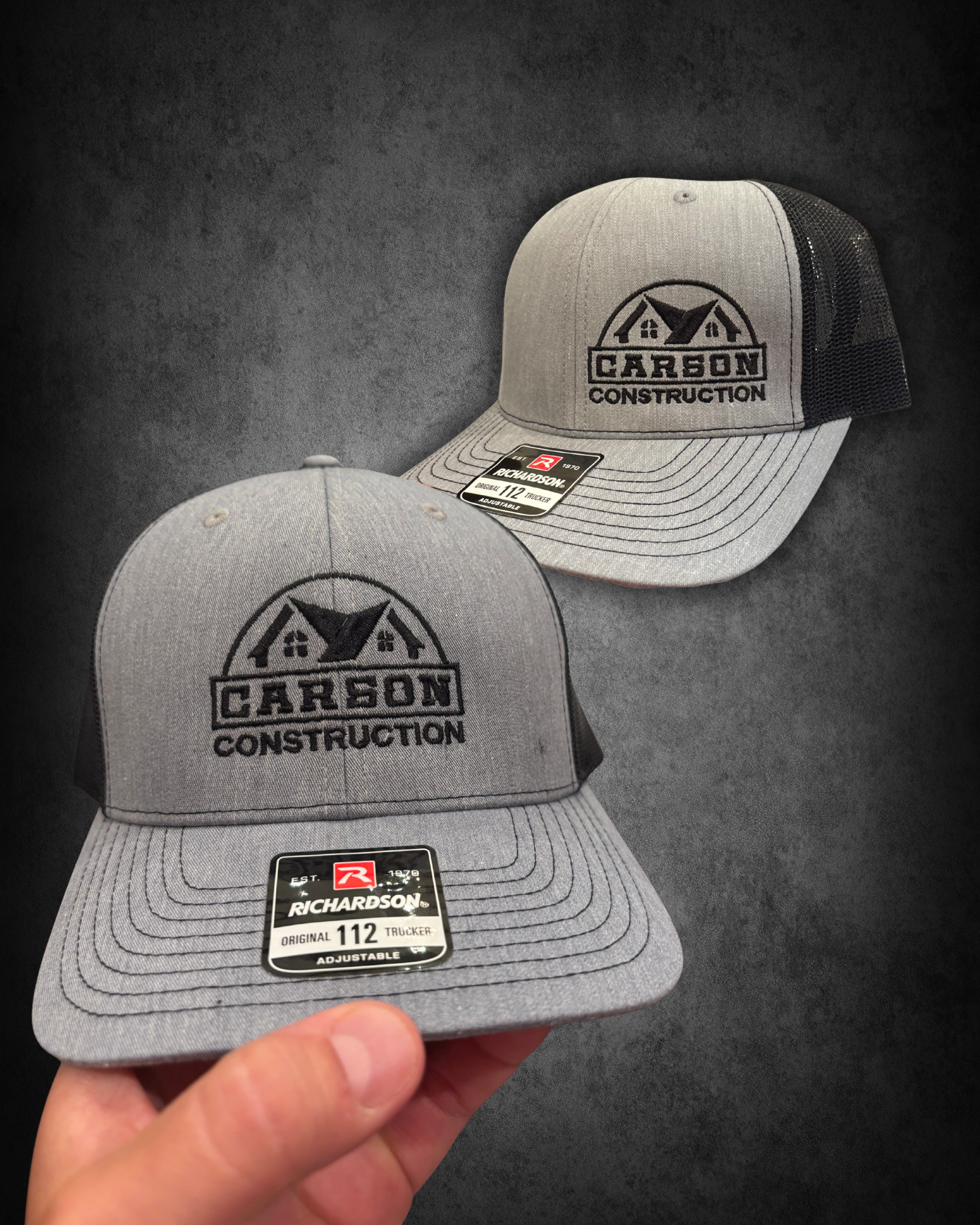 Two gray Richardson trucker caps with black mesh back and front embroidered with 'Carson Construction' logo; one held in hand, one resting on dark surface; stickers on brims.
