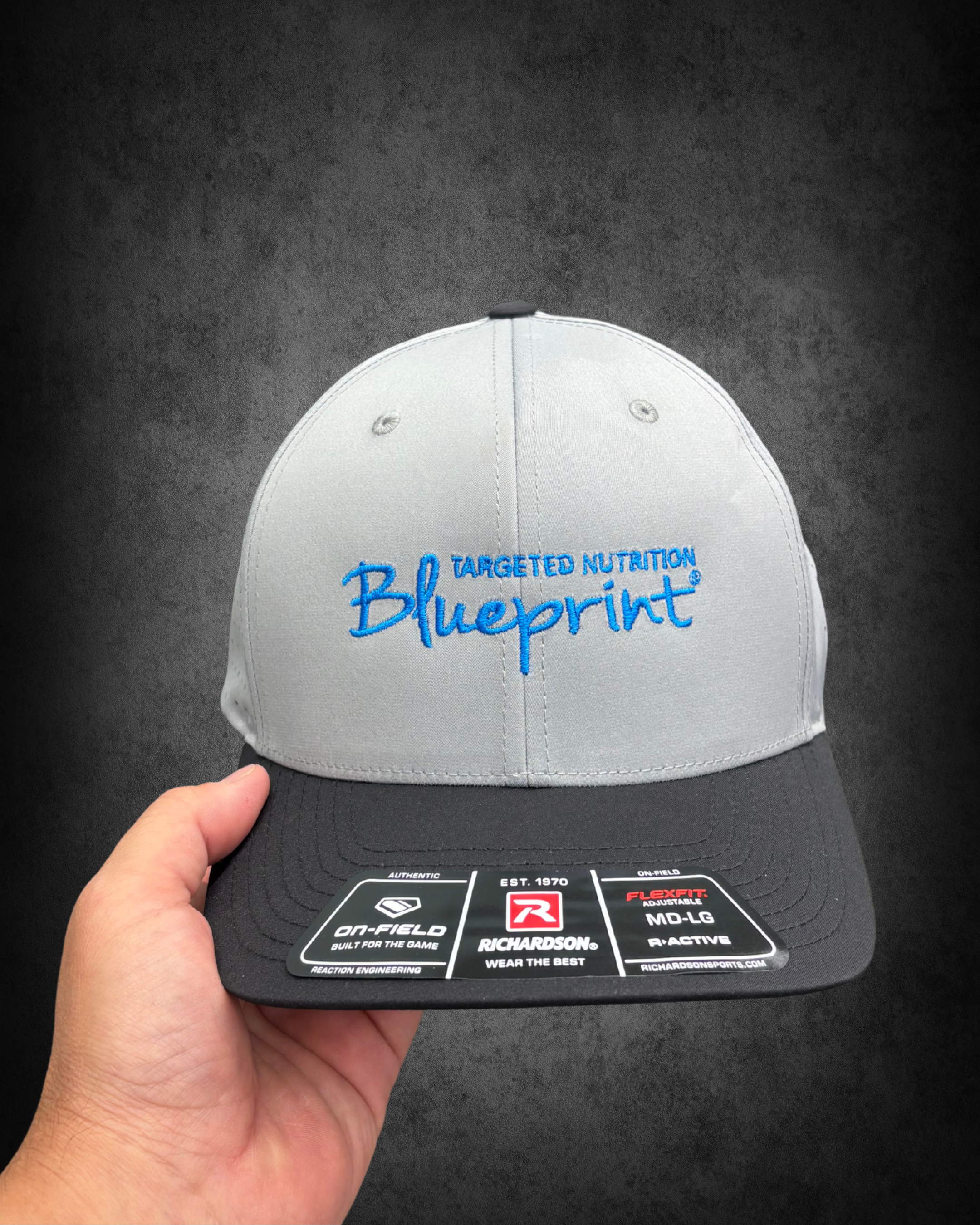 Close-up view of a white baseball cap with blue embroidery reading 'Blueprint' and additional words. A person is holding the cap, and the background is dark.