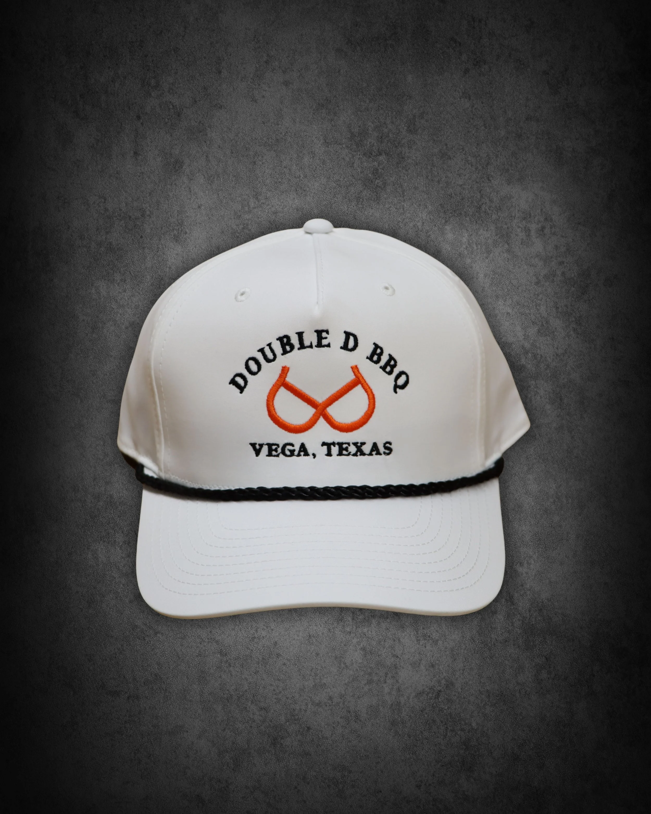 White baseball cap with embroidered logo reading 'Double D BBQ Vega, Texas' and orange barbecue grill graphic on front, placed on a dark surface.