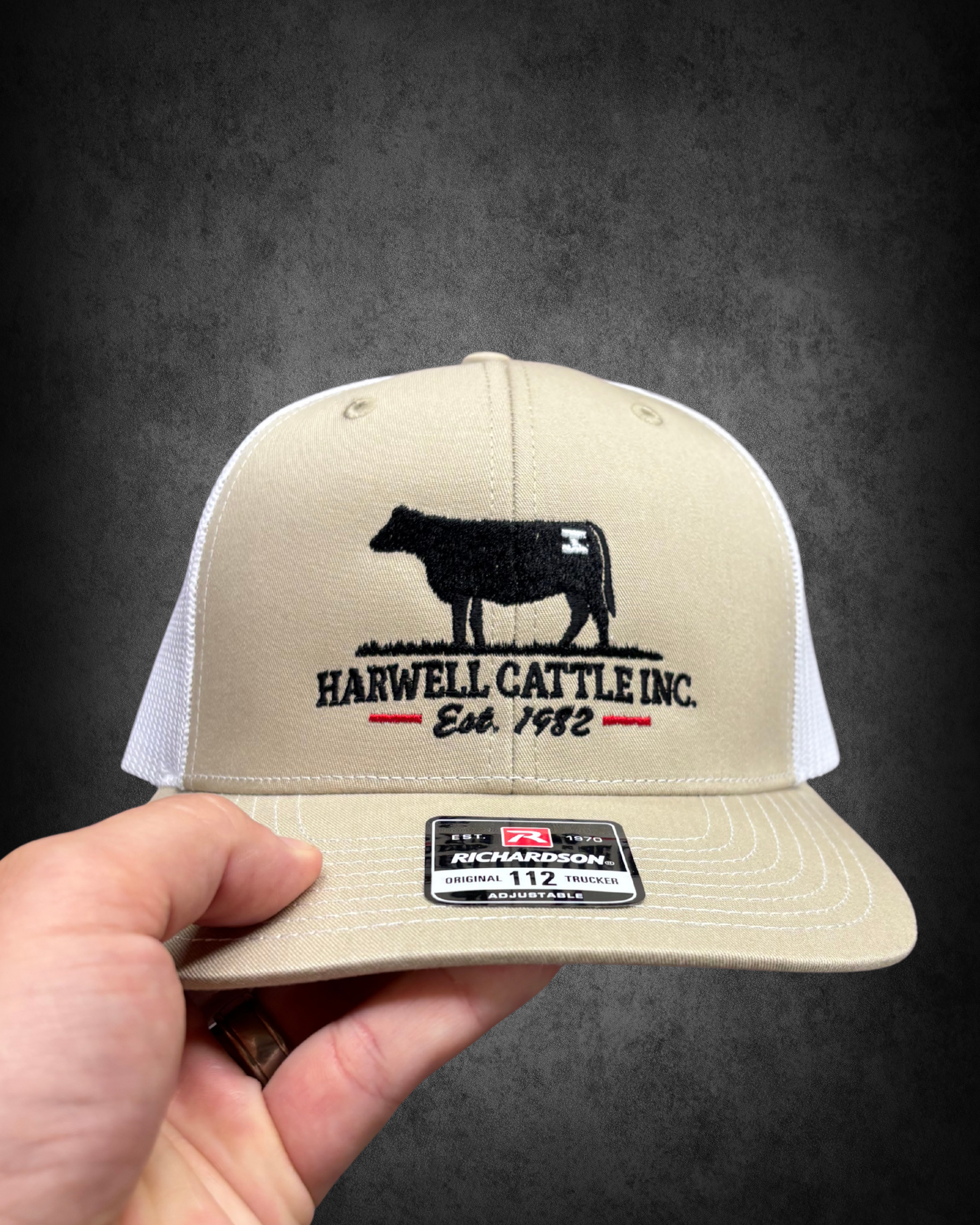 A beige baseball cap with an embroidered black dog silhouette and the text 'HARWELL CATTLE INC. Est. 1982'. A hand is holding the cap, showing the front view. The cap has a Richardson brand sticker on the brim, displaying the size 112 and adjustable 