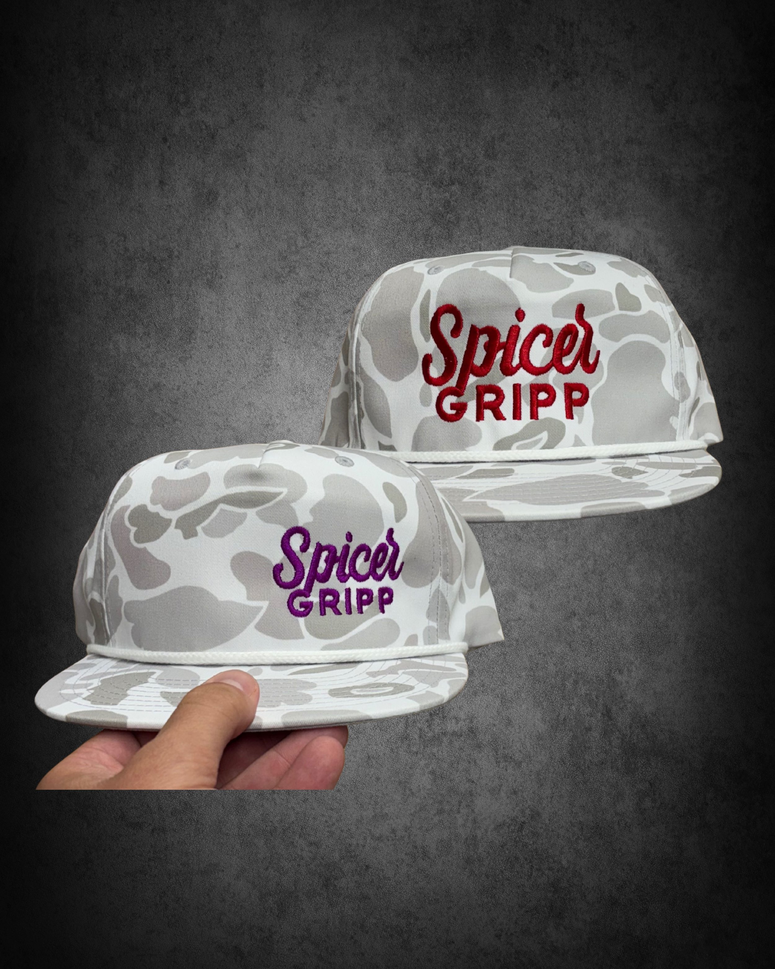 Two white hats with gray abstract patterns, embroidered with the words 'Spice GRIPP' in red and purple, held by a hand against a dark textured background.