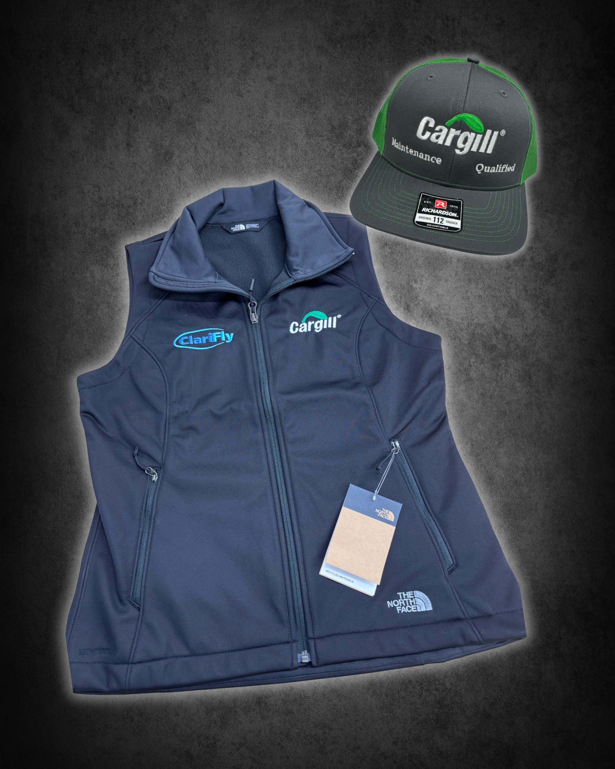 A black and green trucker cap with the brand name 'Cargill' and the words 'Maintenance' and 'Qualified' embroidered on the front. A blue zipped vest with the logos 'Clarify' and 'Cargill' on it, along with a tag, lies below the cap on a dark surface.