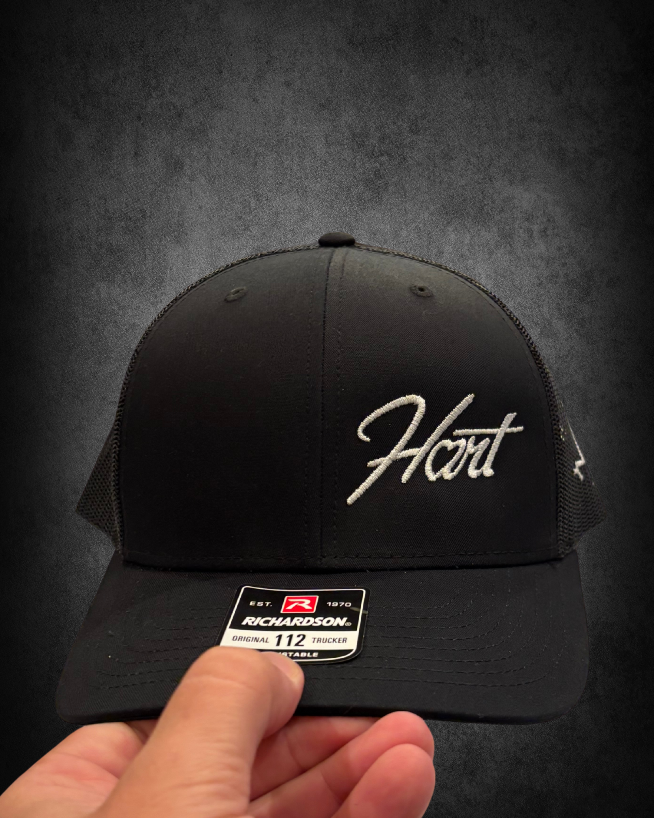 Black baseball cap with embroidered white text, featuring a curved brim and dome shape, on a dark surface. A sticker from Richardson with the brand logo, establishment year, and size is being held by a hand at the front of the cap.