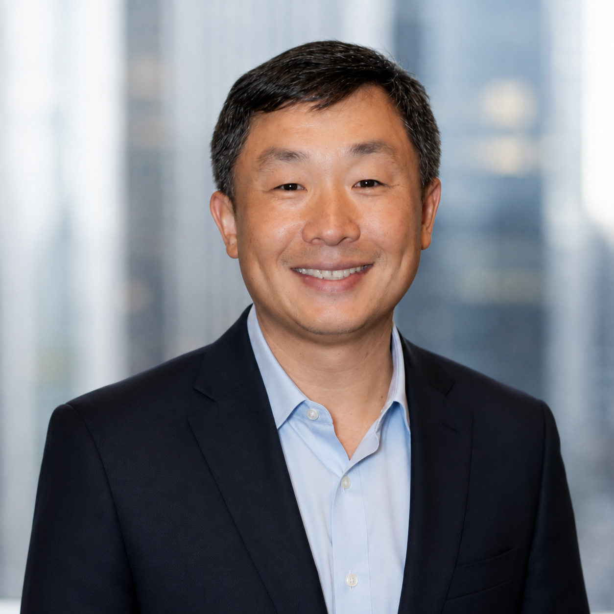 Headshot of an Asian man smiling, wearing a dark suit and light blue shirt, with a blurred modern office background.