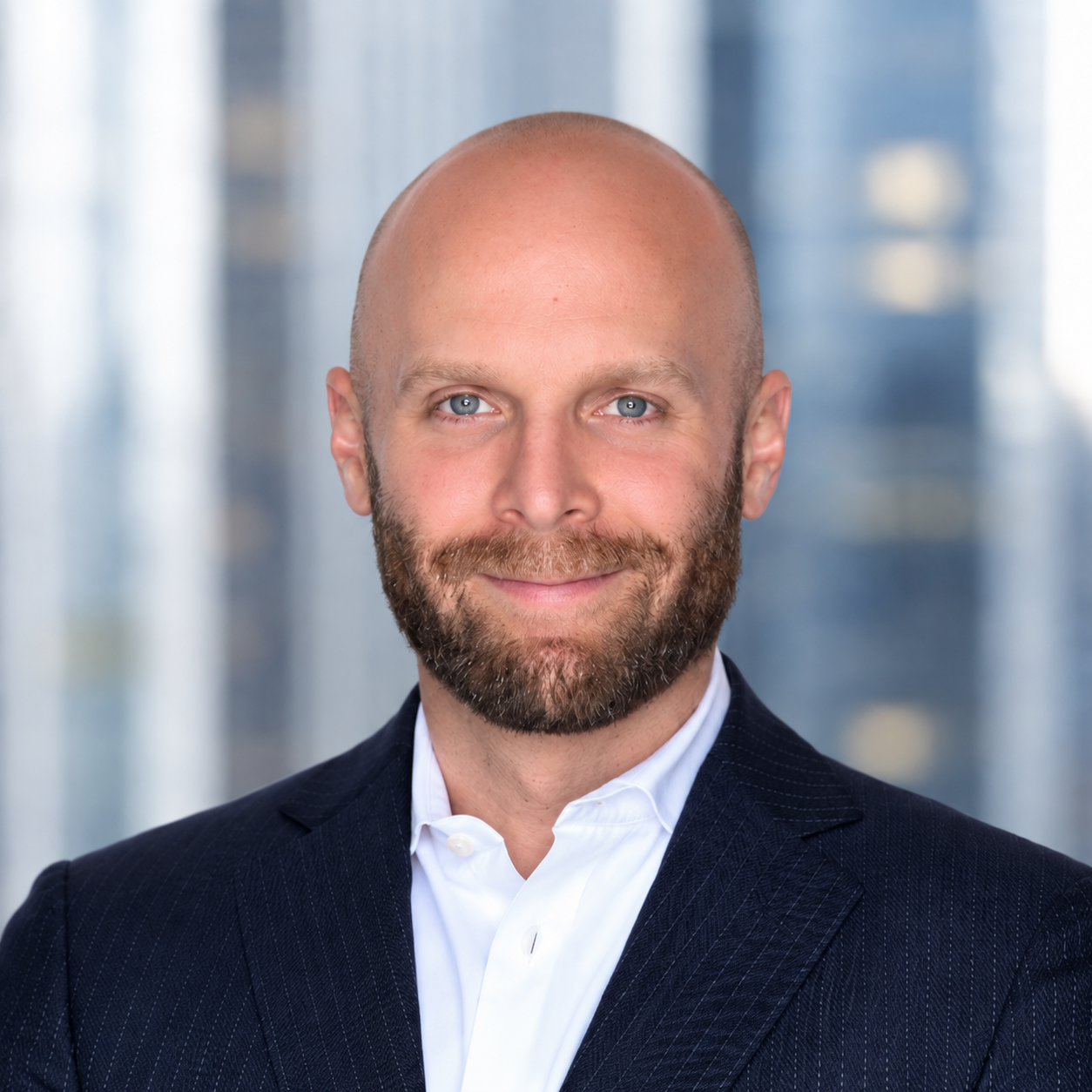 Headshot of a smiling bald man with a beard, wearing a dark suit jacket and white dress shirt, against a blurred office background.